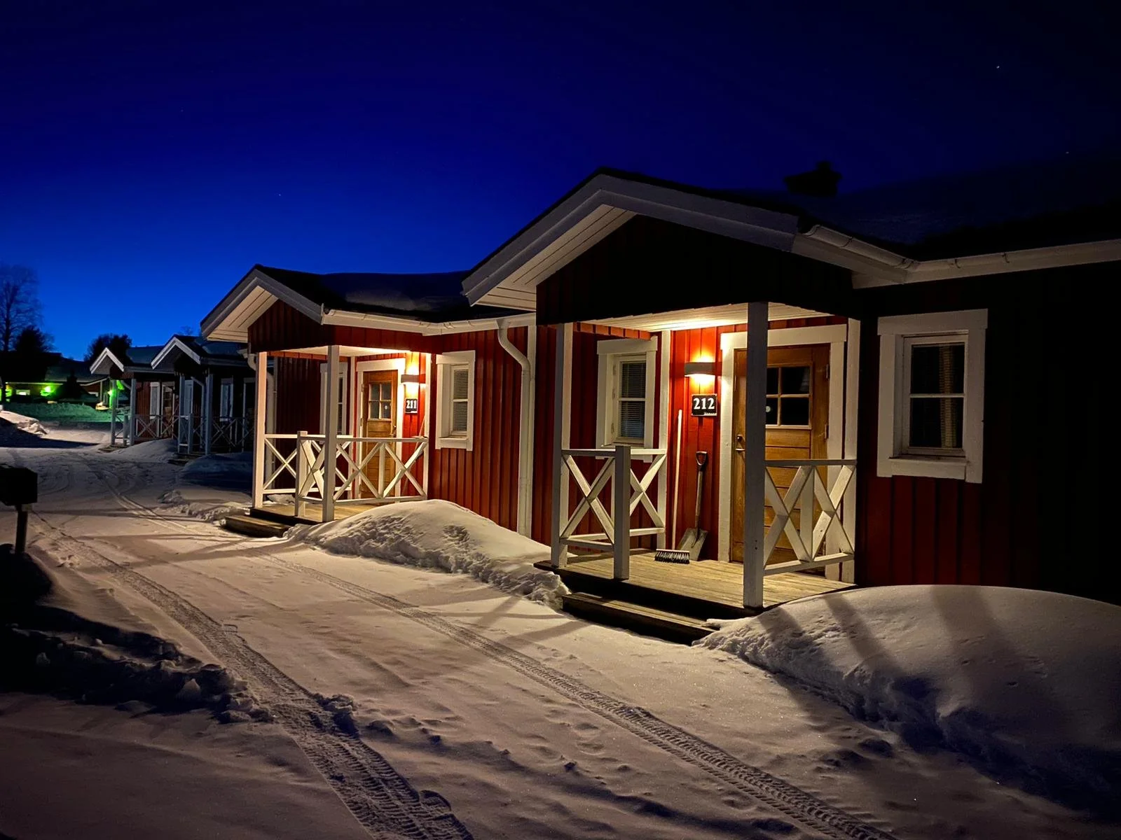 Case di legno rosse con luci accese di notte, neve sul terreno e tracce di pneumatici sulla strada, cielo notturno.