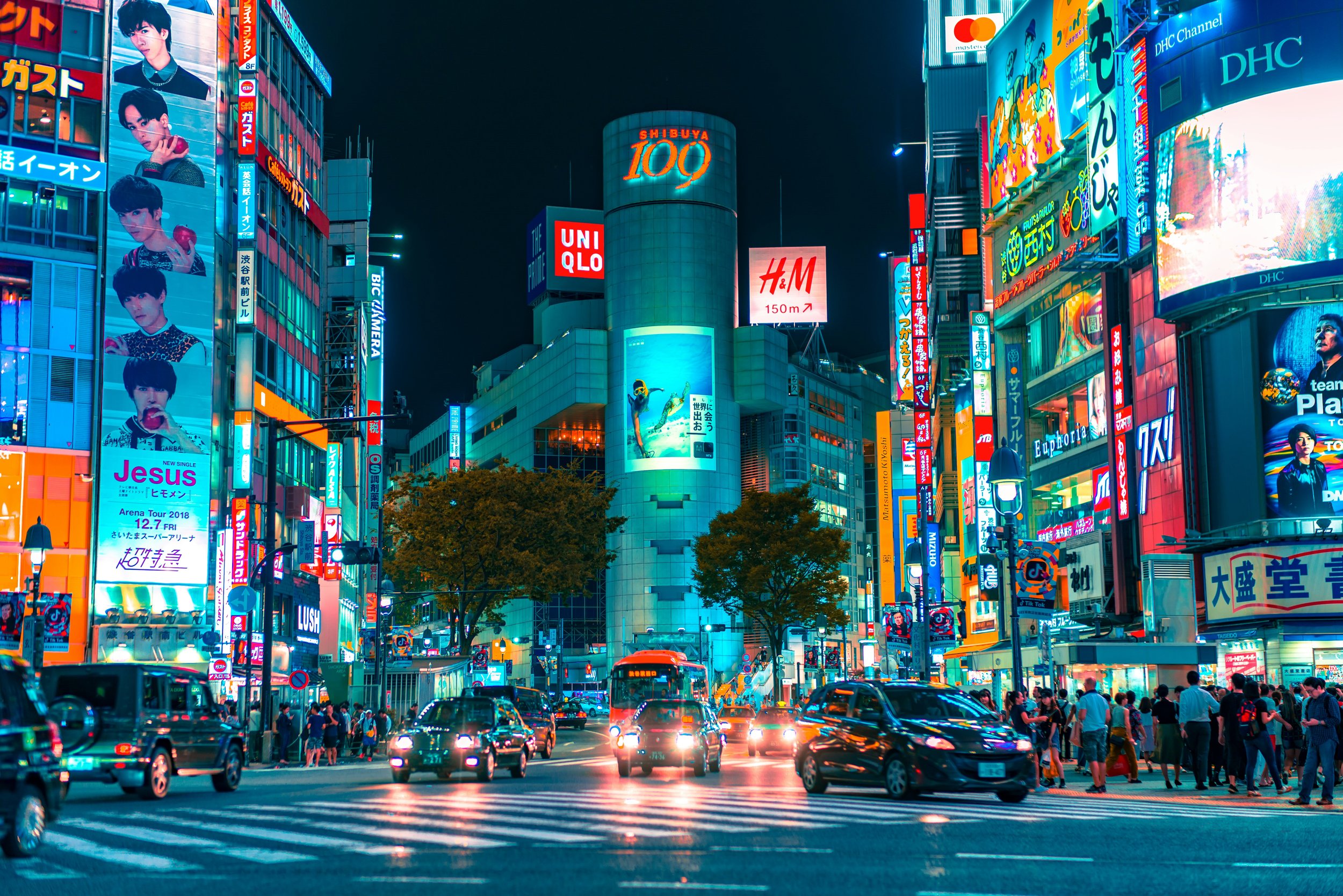 Immagine di una vibrante scena cittadina notturna a Shibuya, Tokyo, con luci al neon, schermi pubblicitari digitali, traffico e pedoni.