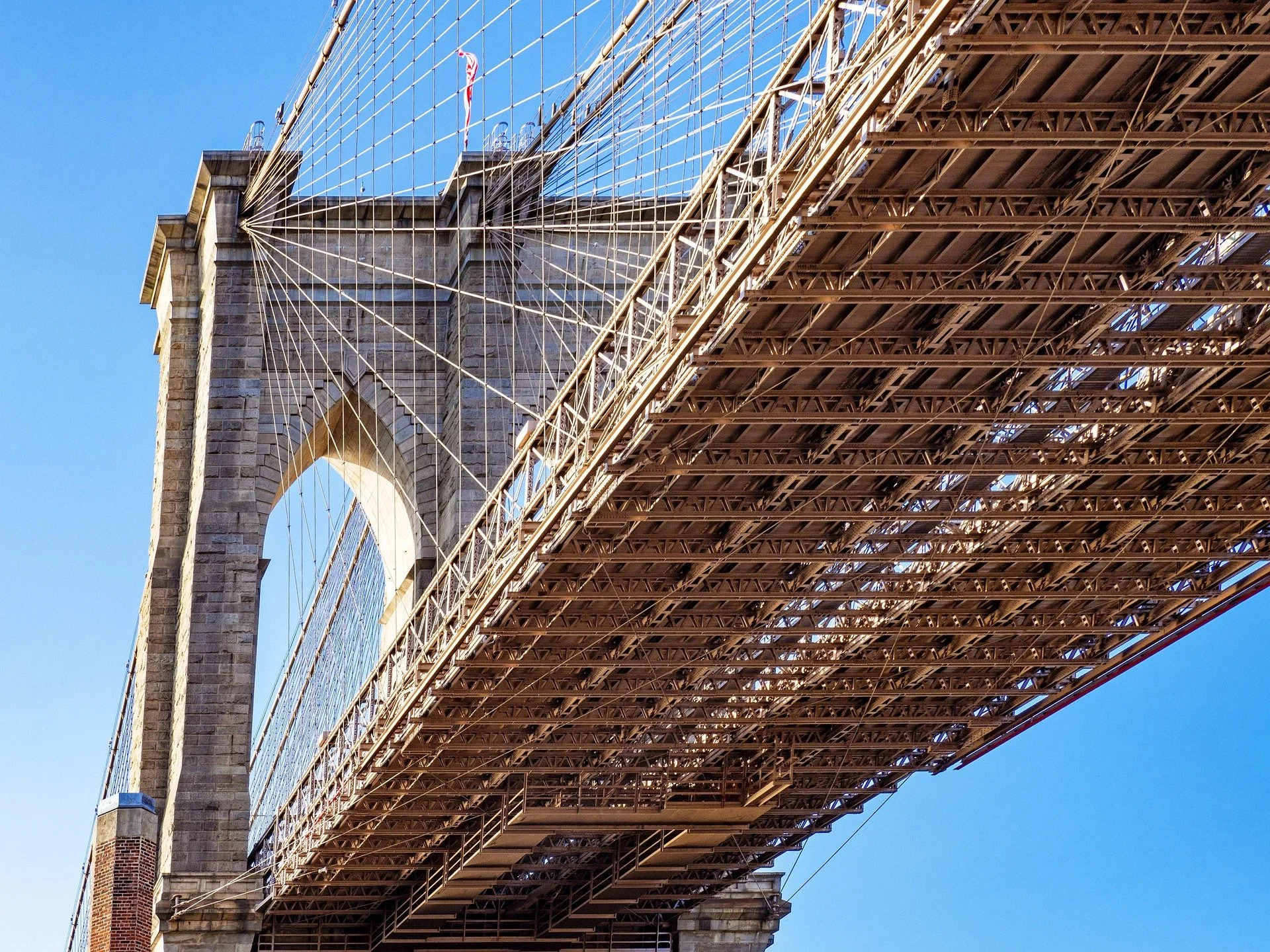 Parte della struttura del ponte di Brooklyn, con le torri in pietra e le sospensioni di cavi, visibile sotto il cielo blu.