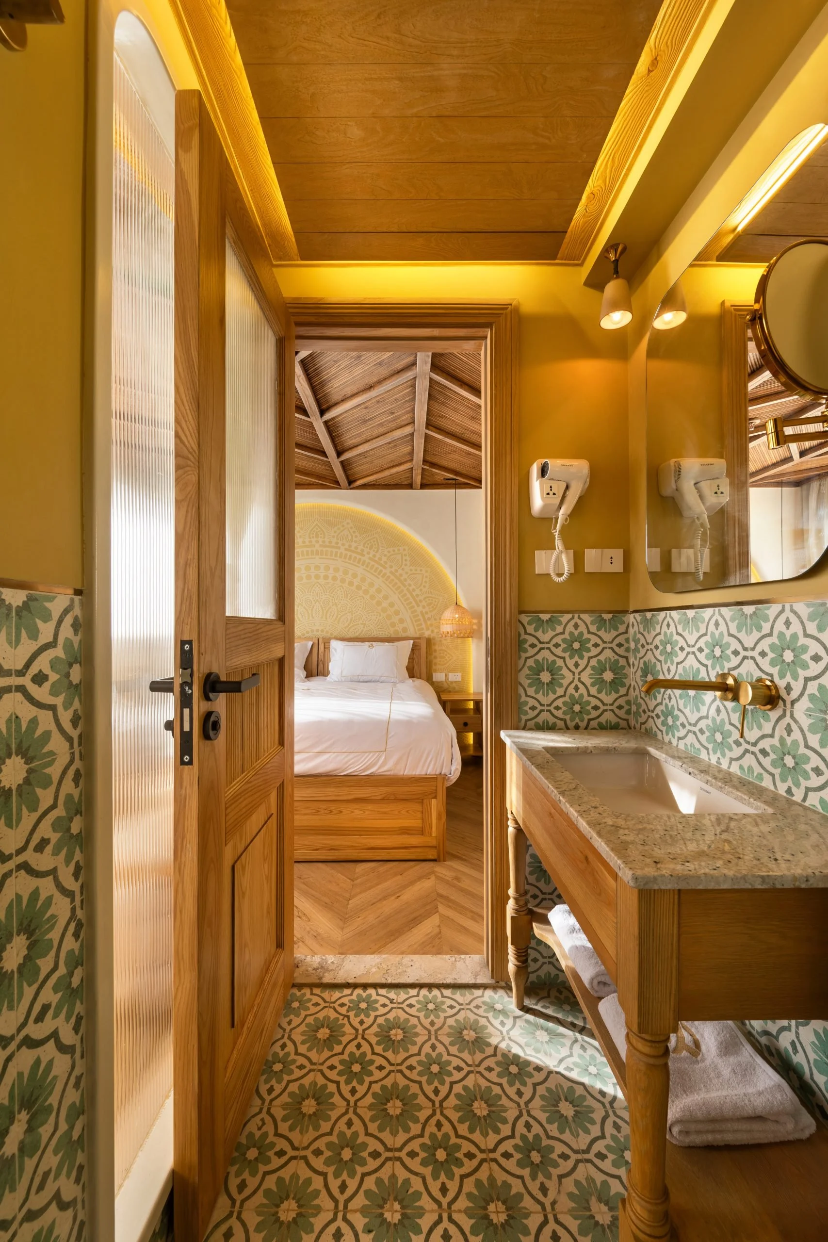 Vista di una camera da letto attraverso una porta aperta, si vede un letto con lenzuola bianche, un lavabo in marmo con asciugamani piegati sotto, pareti decorate con piastrelle verdi e un soffitto di legno.