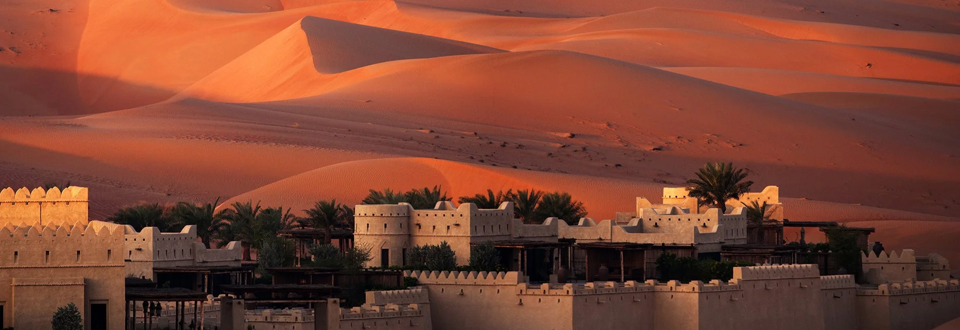 Una fortezza o villaggio nel deserto con mura di argilla e palme, sullo sfondo dune di sabbia rosa