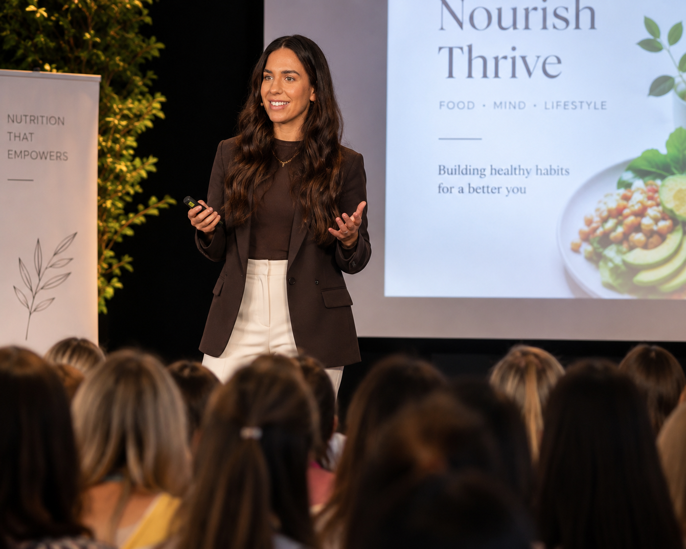 A woman speaking at a presentation with a large screen behind her displaying the title 'Nourish Thrive' and a subtitle about building healthy habits, with an audience of several people visible in the foreground.