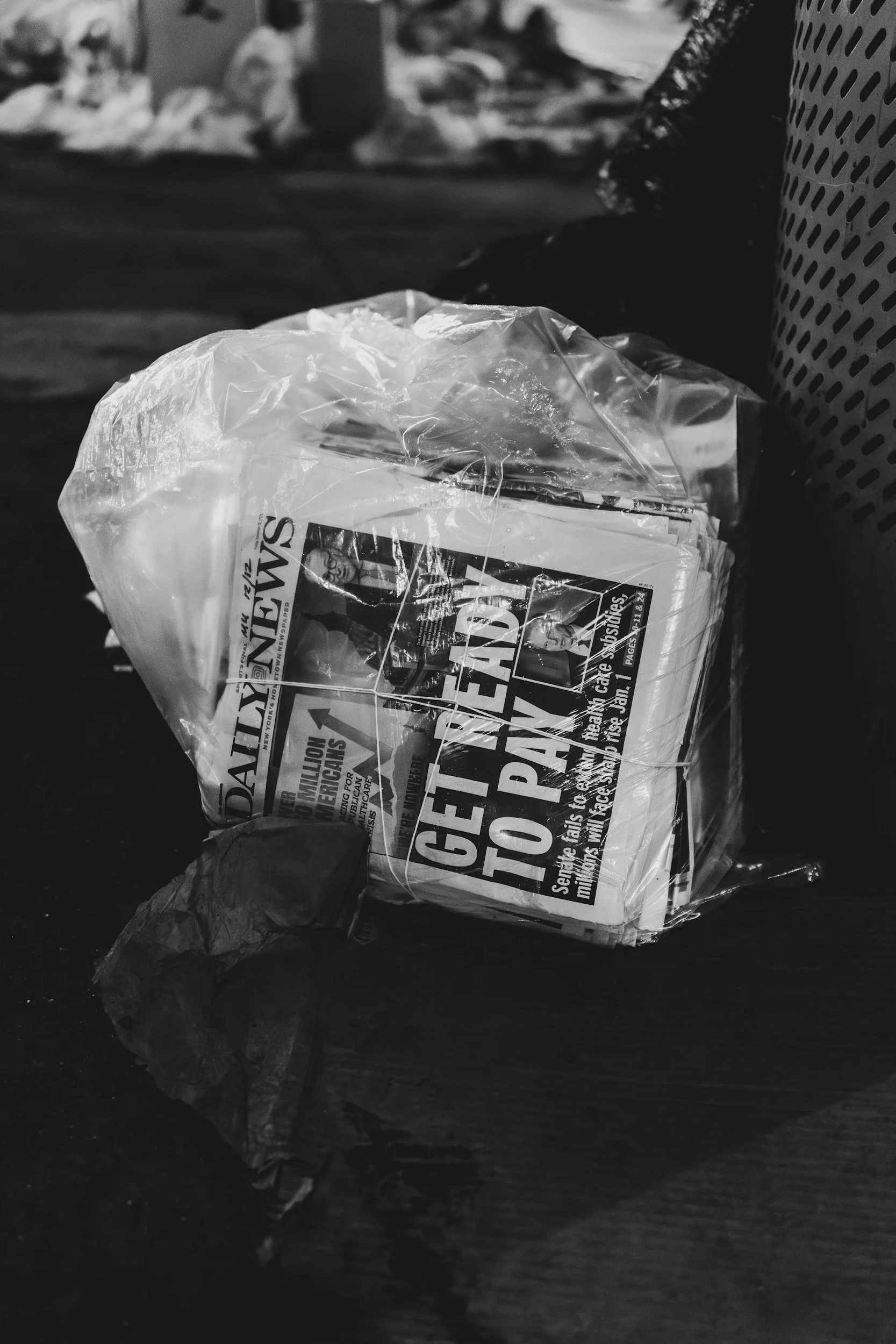A crumpled plastic bag containing newspapers and trash lying on a dark wooden surface, with more garbage in the blurred background.
