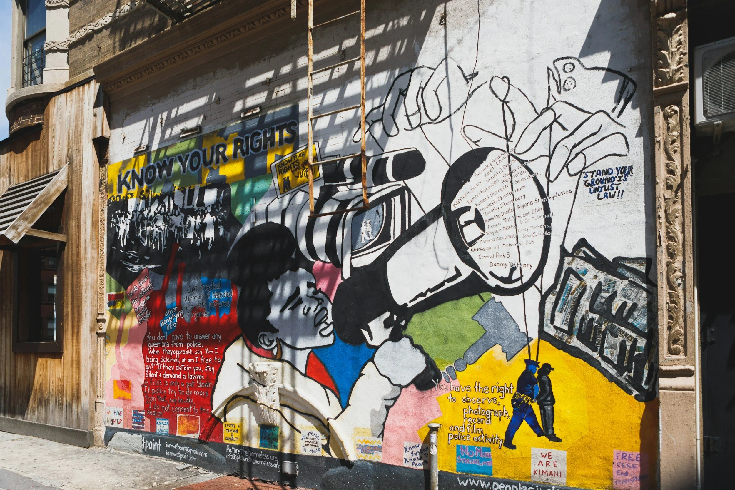 A colorful mural on an urban brick wall depicting two children talking to a police officer, with various messages about rights, law, and activism written around the artwork. The mural includes illustrations of hands, a camera, and people in protest.