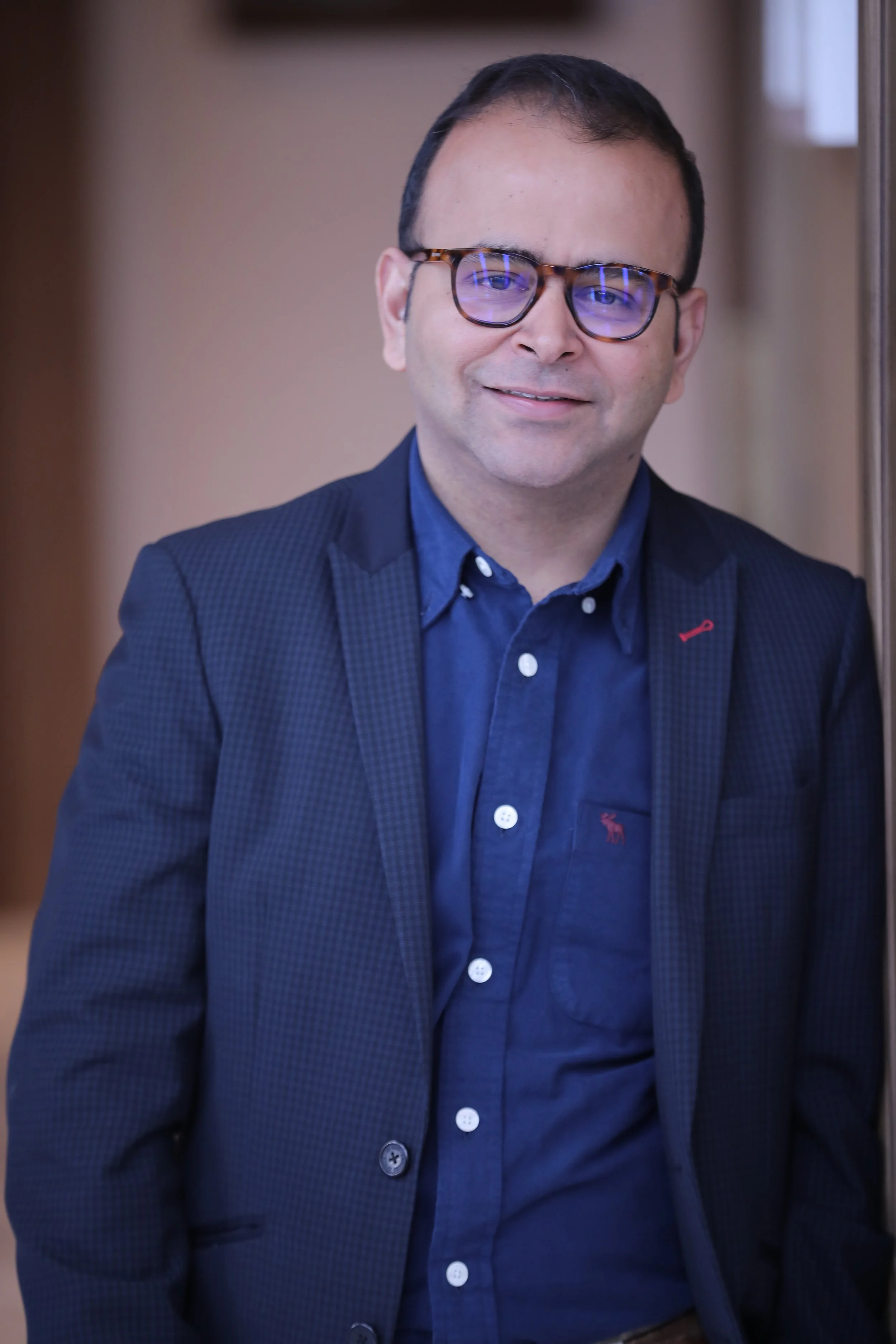 A man with short dark hair and glasses wearing a dark blue blazer and shirt, smiling and looking at the camera.