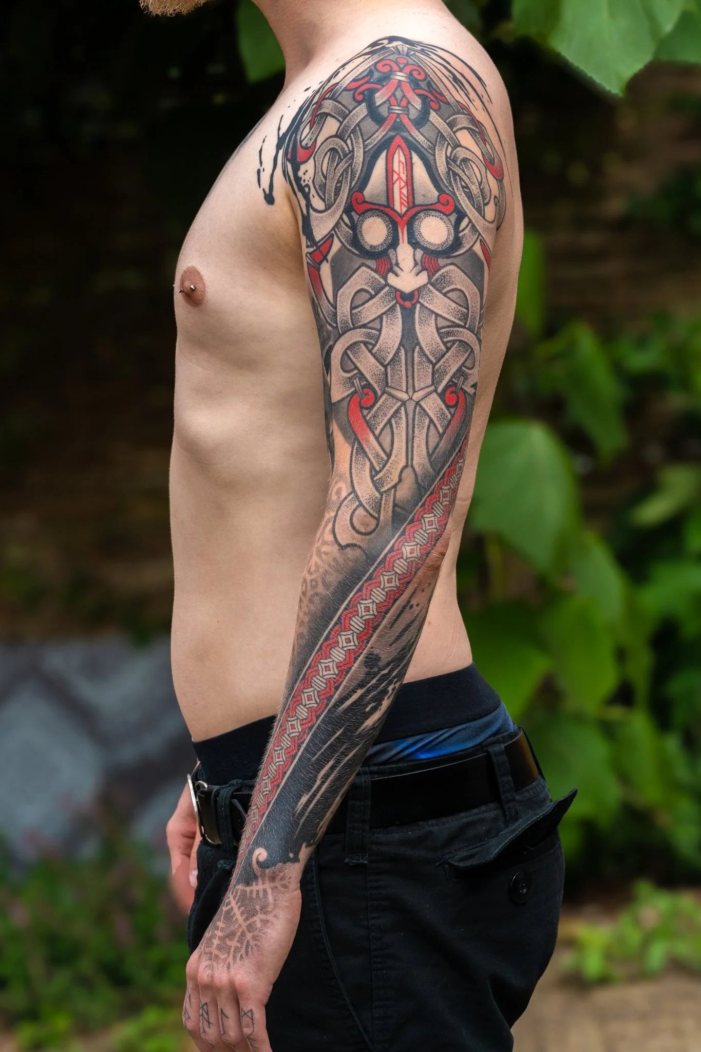 A person with tattoos on their arm and chest, wearing black pants and a belt, standing outdoors amidst green foliage.