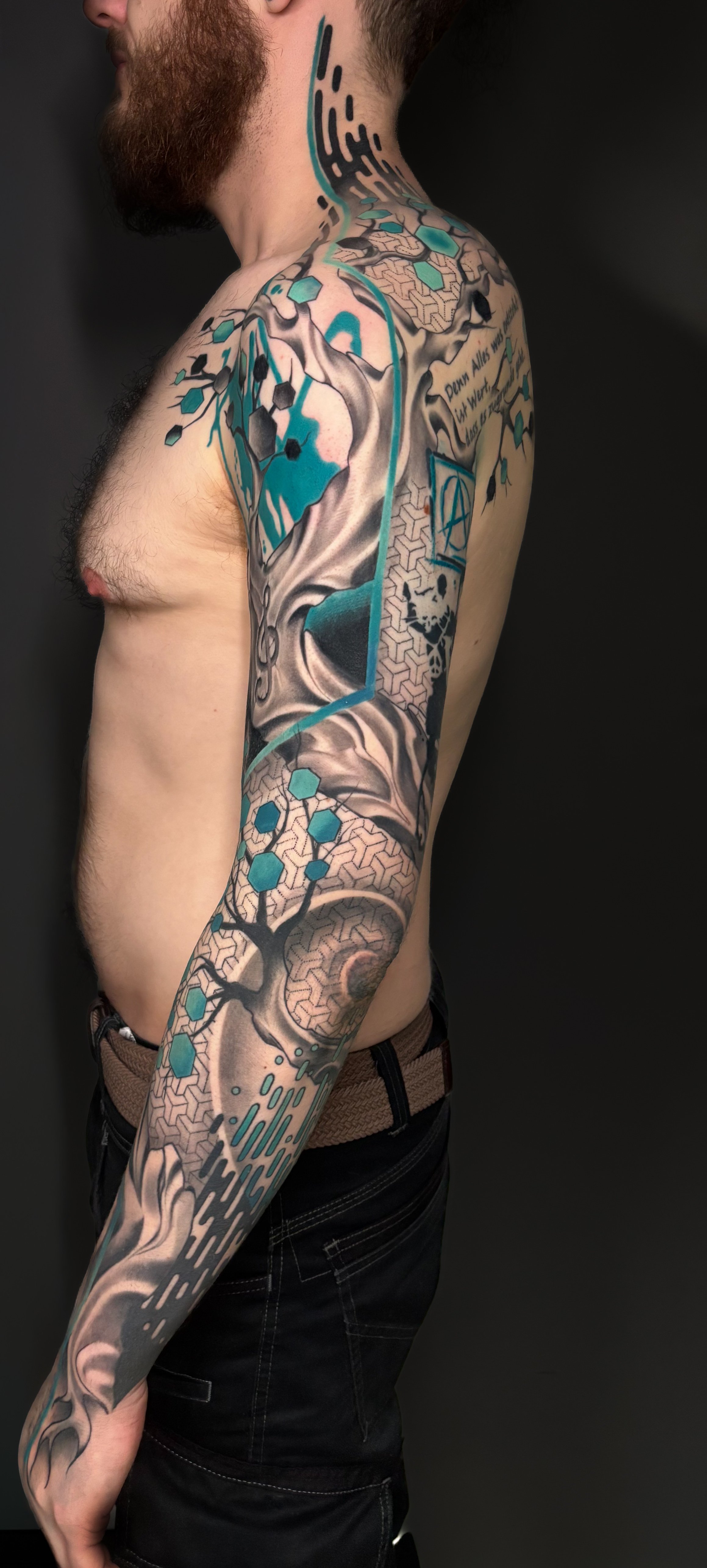 A man's upper body with a sleeve tattoo featuring a tree, geometric patterns, and abstract designs in black, gray, turquoise, and beige tones.