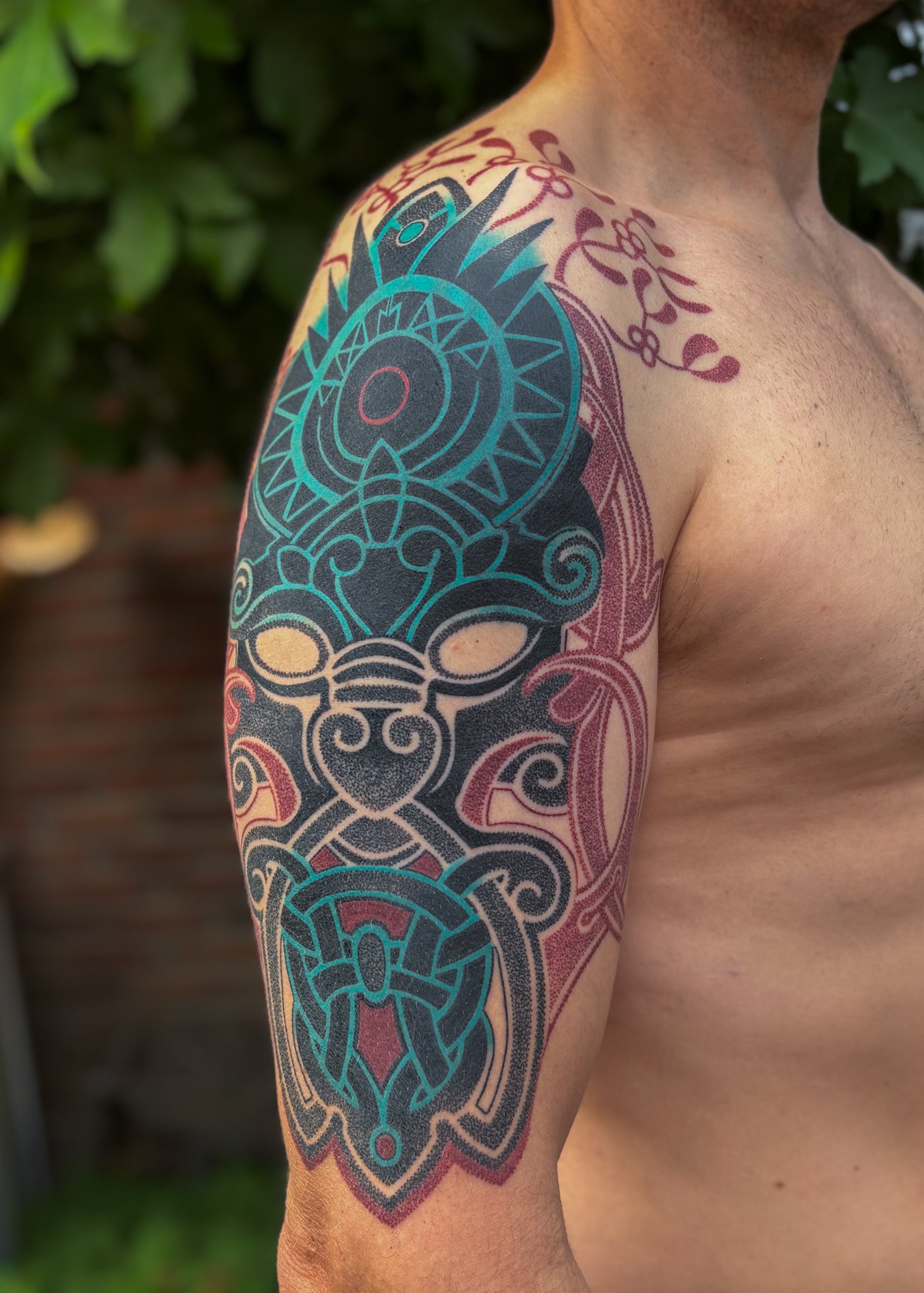 A tattoo on a person's shoulder and upper arm featuring a large, stylized, blue and red tribal or geometric design with intricate patterns.