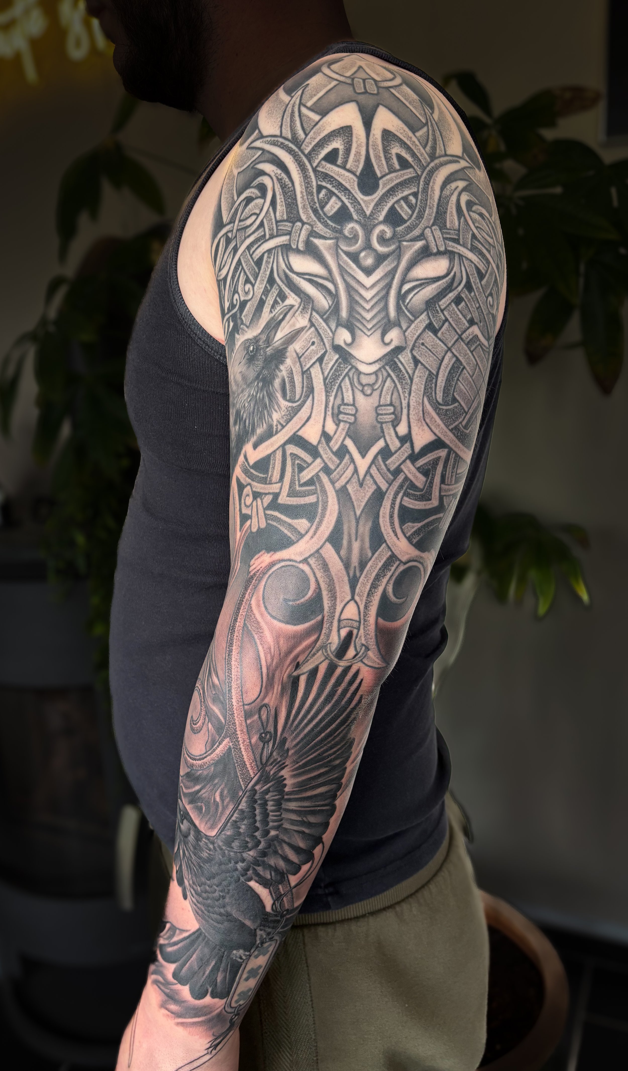 Close-up of a person's arm with detailed black and gray tribal-style tattoo art featuring a wolf, a bird, and abstract patterns.
