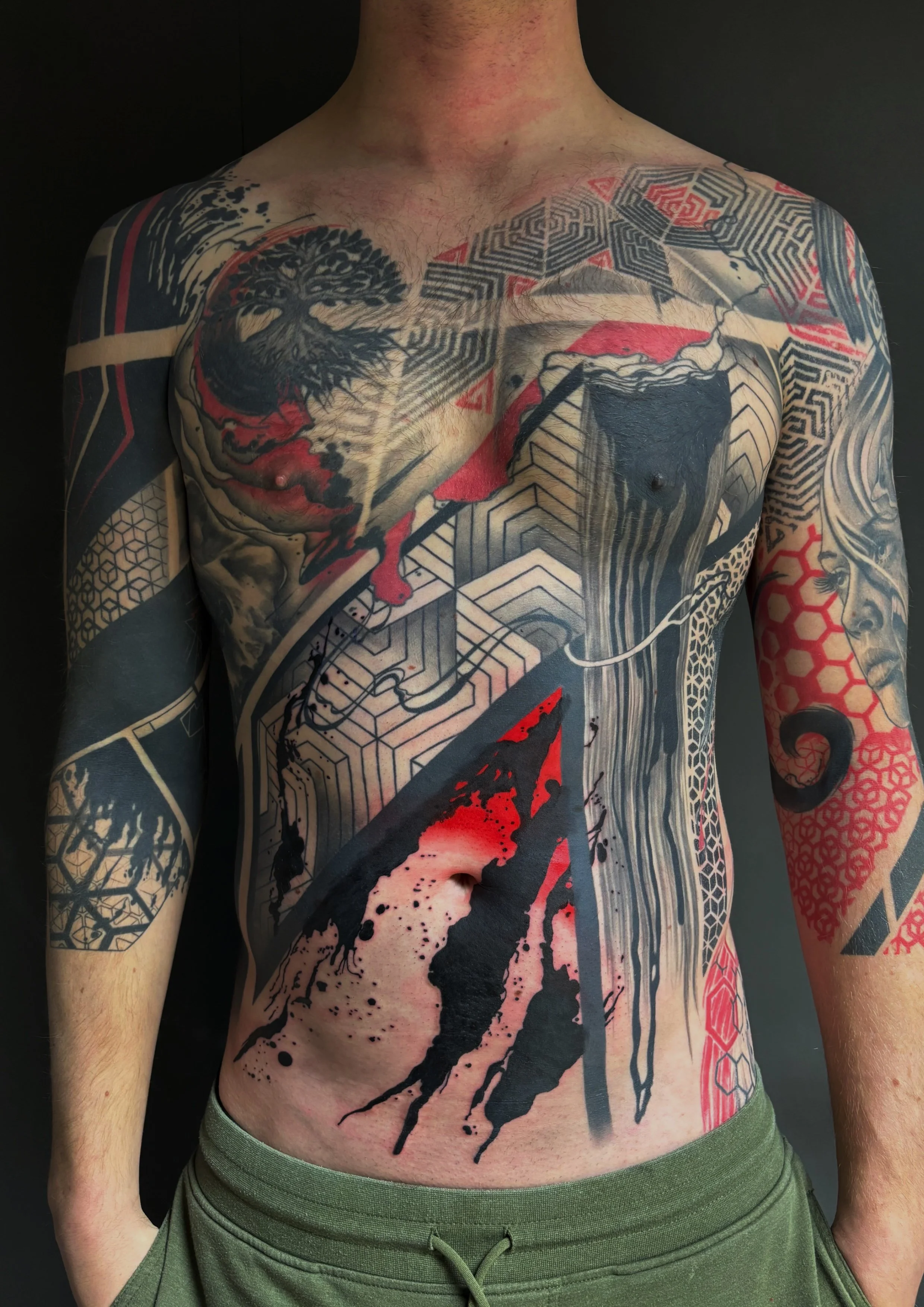 Close-up of a person's torso with extensive tattoos, including a tree, geometric patterns, a red and black abstract design, and a dragon head, against a black background.