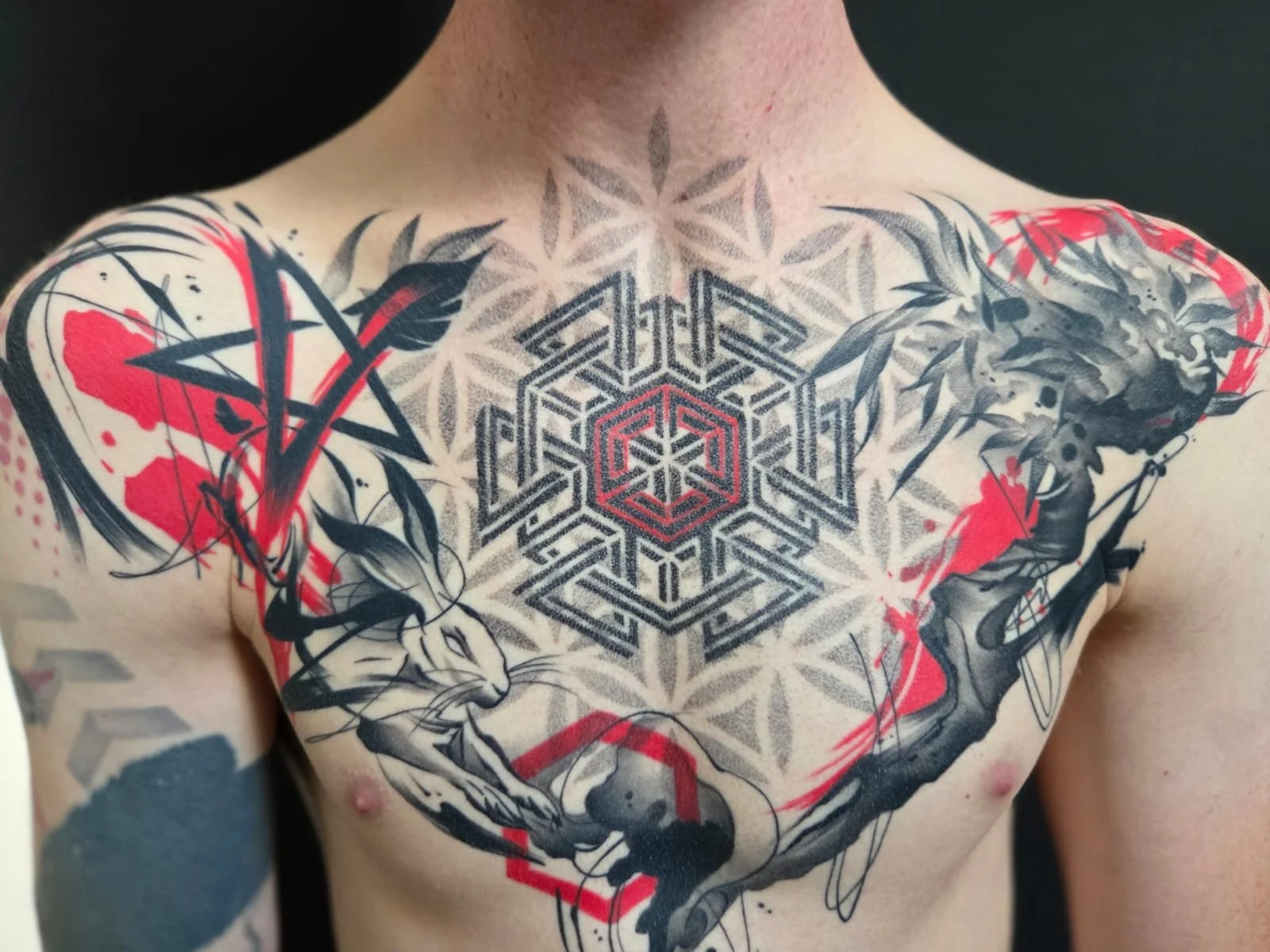 Close-up of a person's chest showing an intricate tattoo design with geometric patterns, a stylized horse, and abstract elements in black, red, and gray ink.