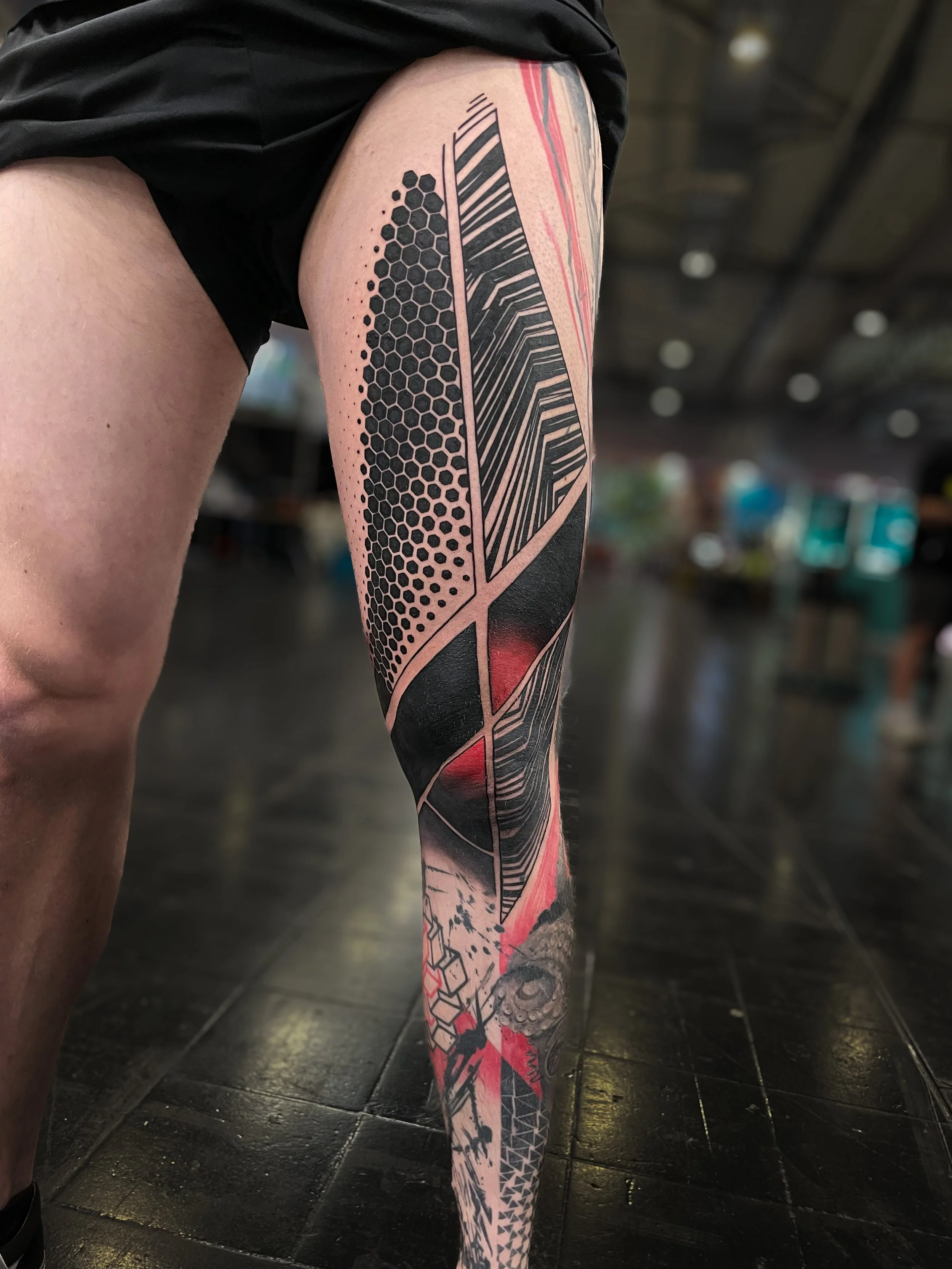 Close-up of a person's leg with a colorful geometric and abstract tattoo, standing on a dark tiled floor in an indoor setting.