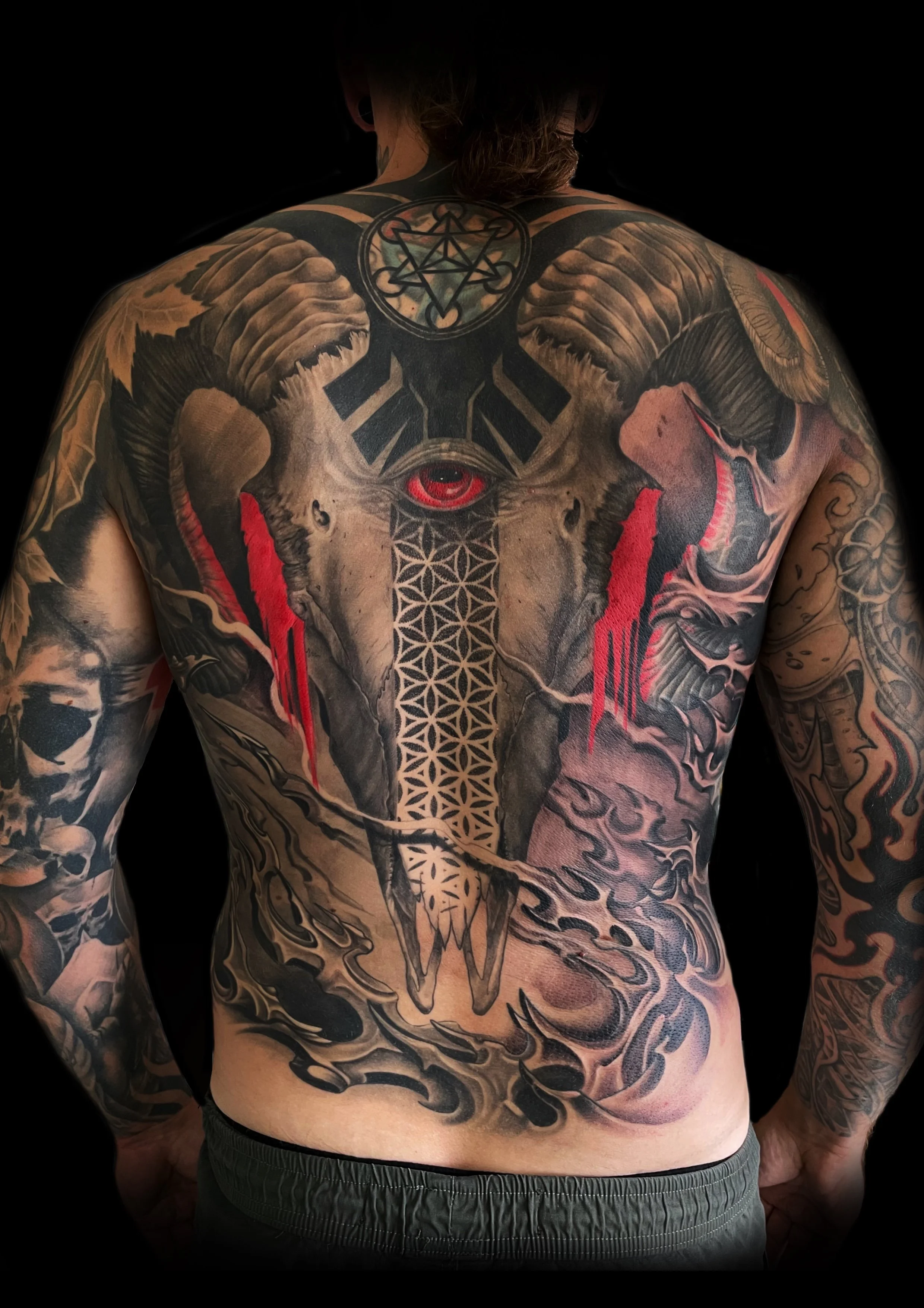 Tattooed man with a Ram's head tattoo in the center of his chest, featuring large horns, an eye, geometric patterns, and red accents, with additional tattoos on his arms and shoulders.