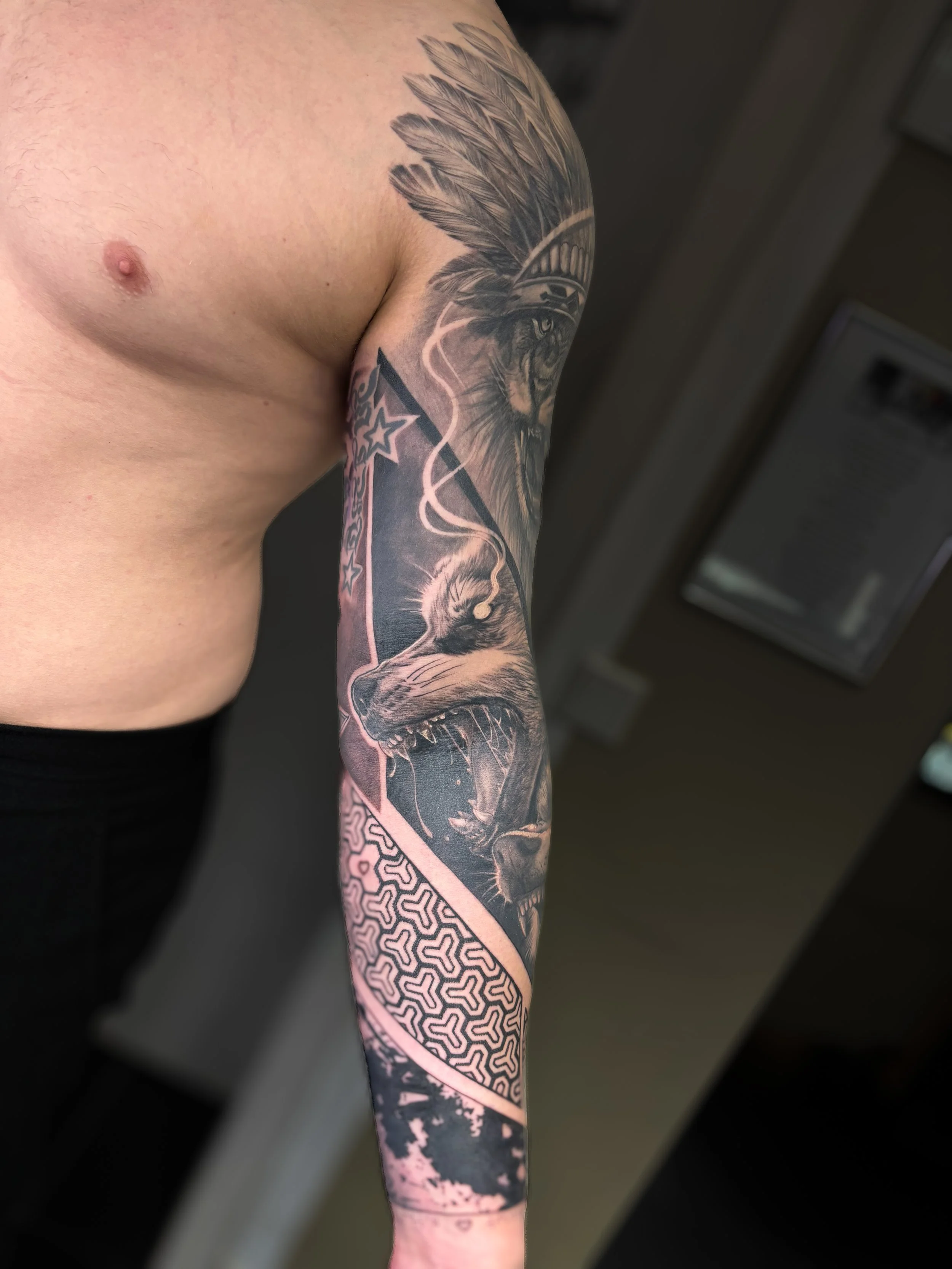 Close-up of a person's upper arm and shoulder with a large, detailed black and gray tattoo of a lion with a feathered headdress, and a geometric pattern on the forearm.