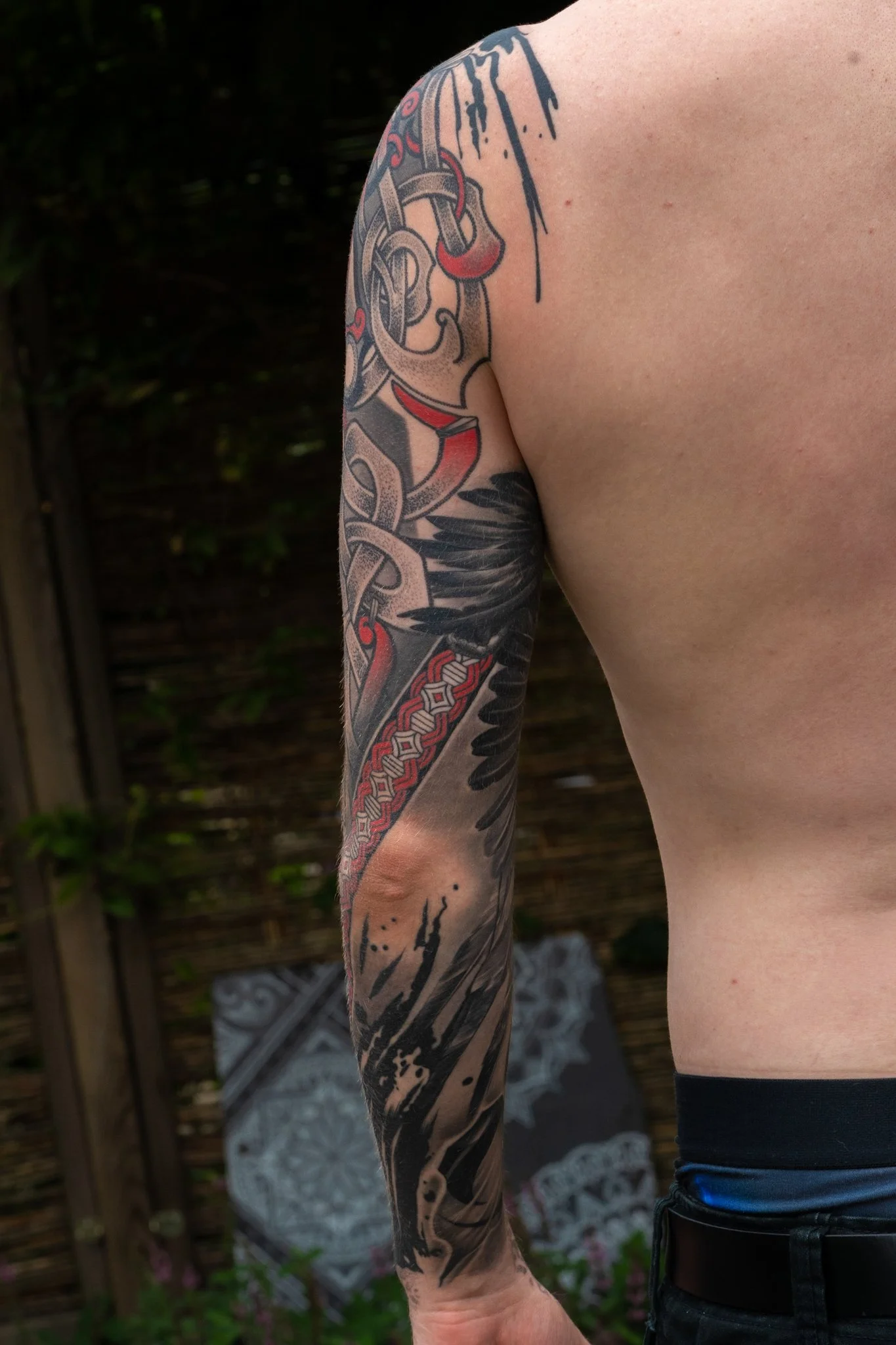 Close-up view of a person's arm featuring a detailed, colorful tattoo with black, red, and gray elements extending from the shoulder to the wrist. The person is shirtless, standing outdoors with a wooden fence and some greenery in the background.