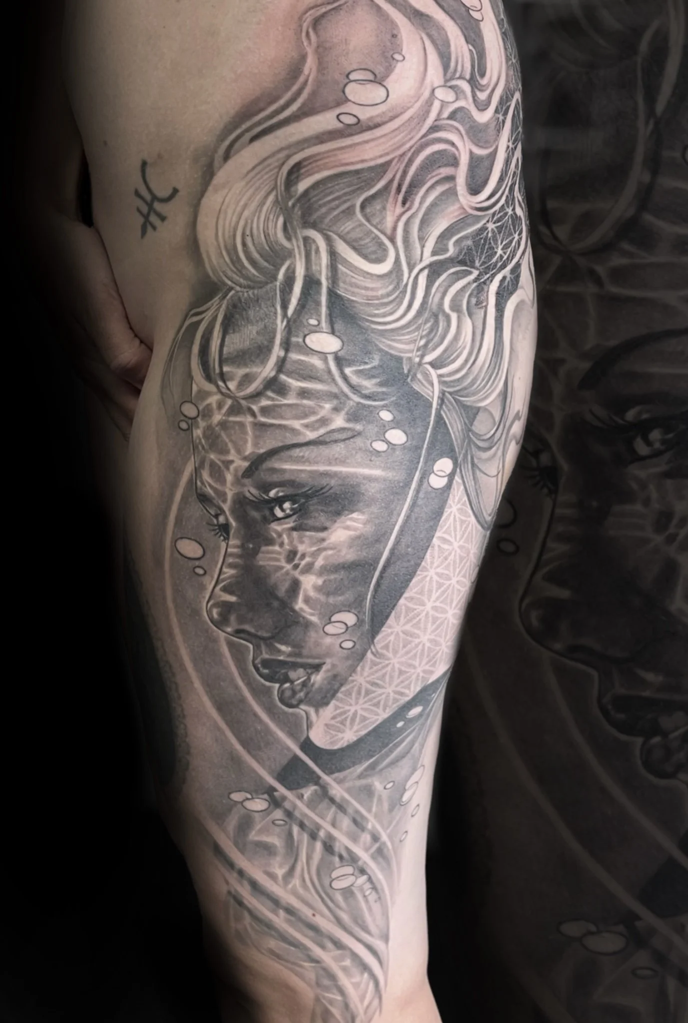 Close-up of a person's arm with a detailed black and gray tattoo design featuring a face, flowing hair, and abstract geometrical elements.