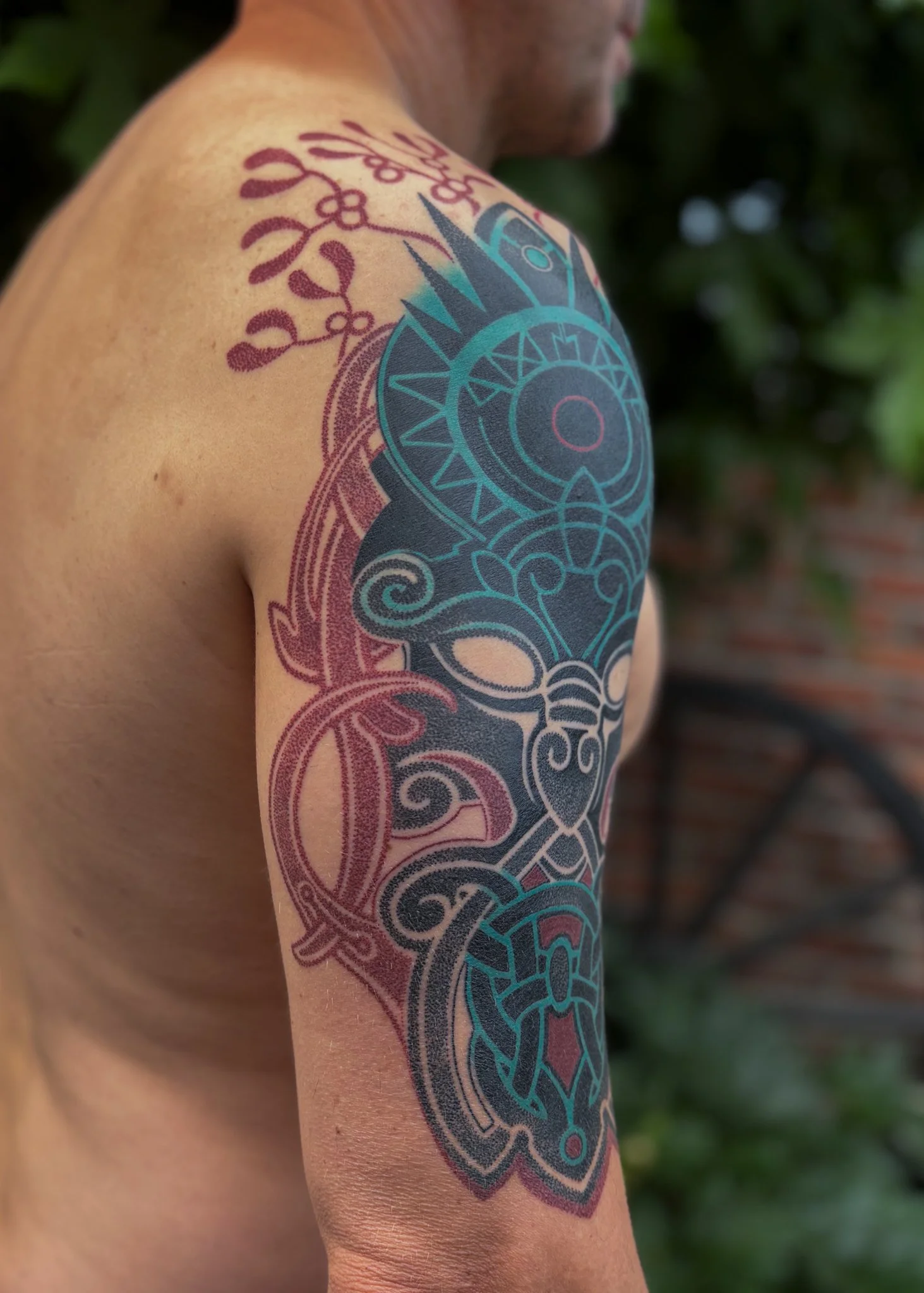 Close-up of a person's shoulder and upper arm with a colorful tribal-style tattoo design that includes geometric patterns and floral elements. Background of greenery.