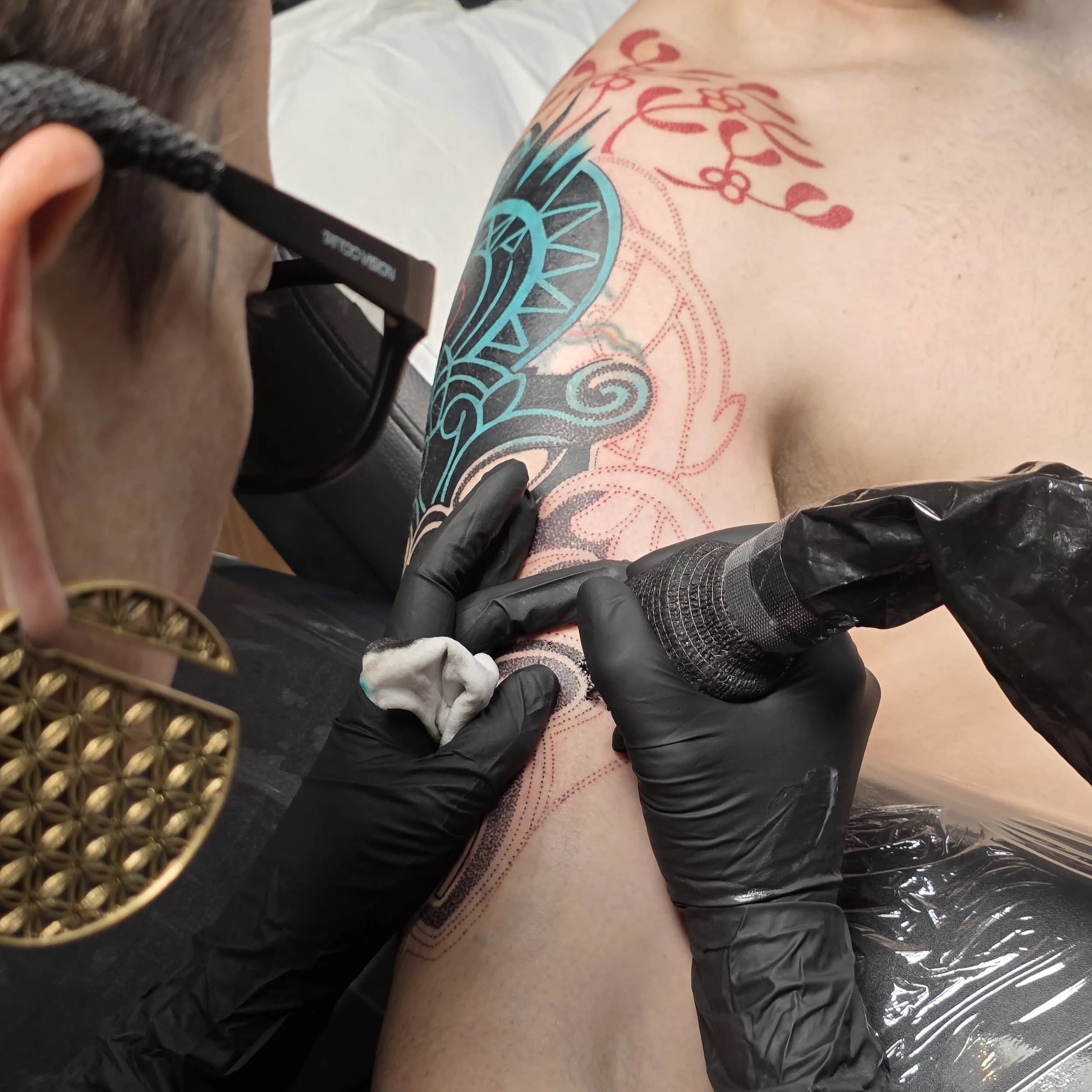 A tattoo artist is tattooing a colorful design on a person's leg. The person is lying down on a surface covered with plastic wrap, and the tattoo incorporates blue, black, and red ink with intricate patterns. The artist is wearing black gloves and holding a tattoo machine.