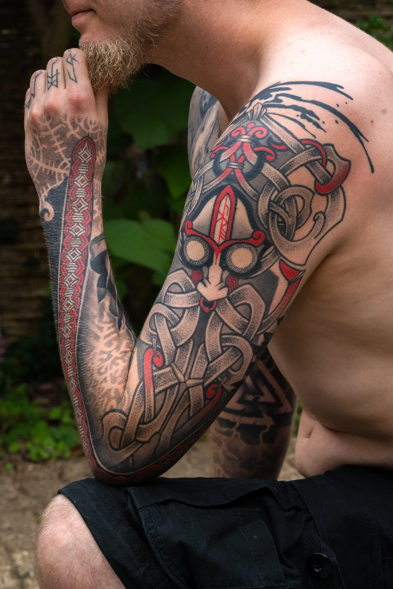 A close-up of a shirtless man with tattoos on his arm and chest, sitting outdoors with greenery in the background.