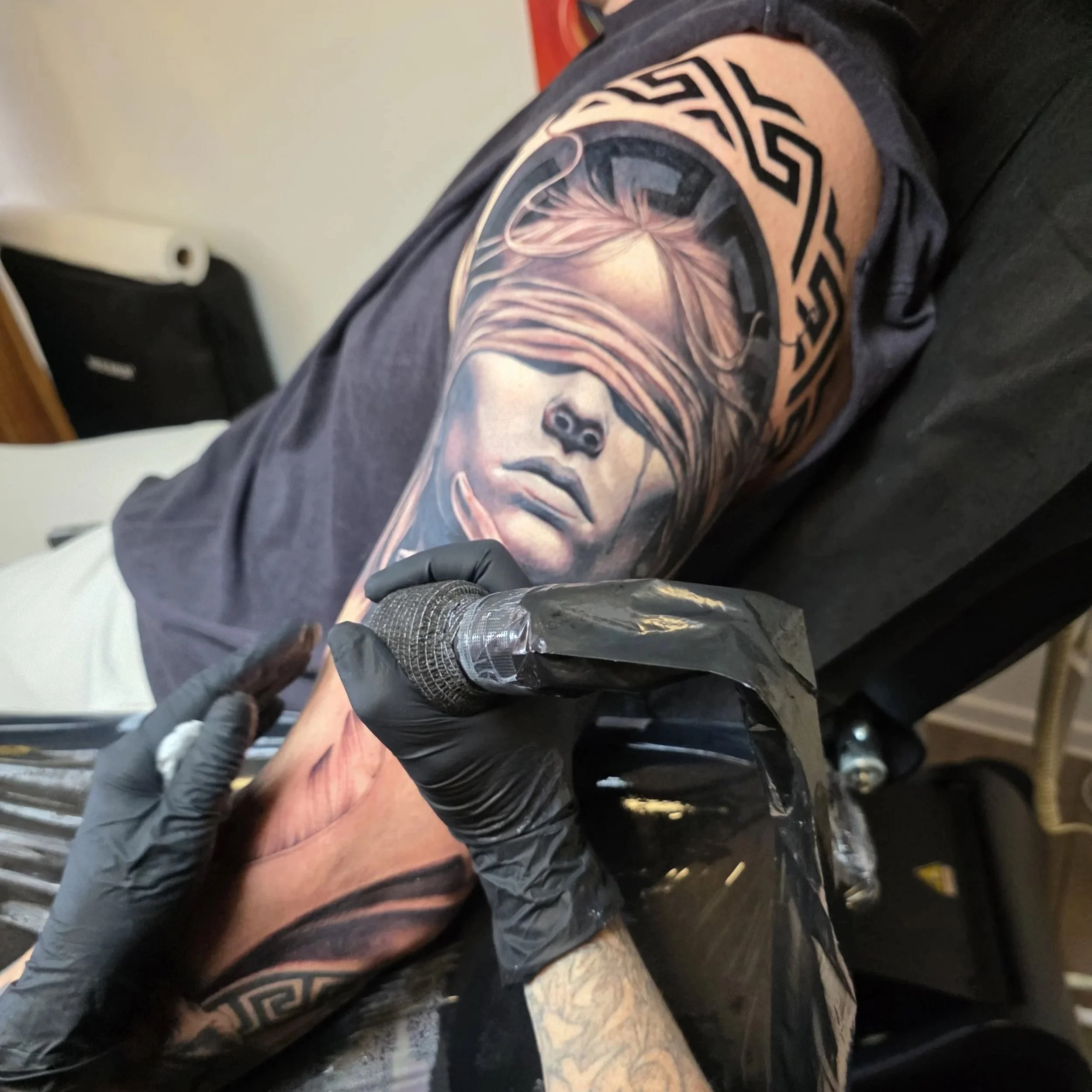 A person with tattoos on their arm, wearing black gloves, holding a tattoo machine, in the process of tattooing a face on another person's skin.