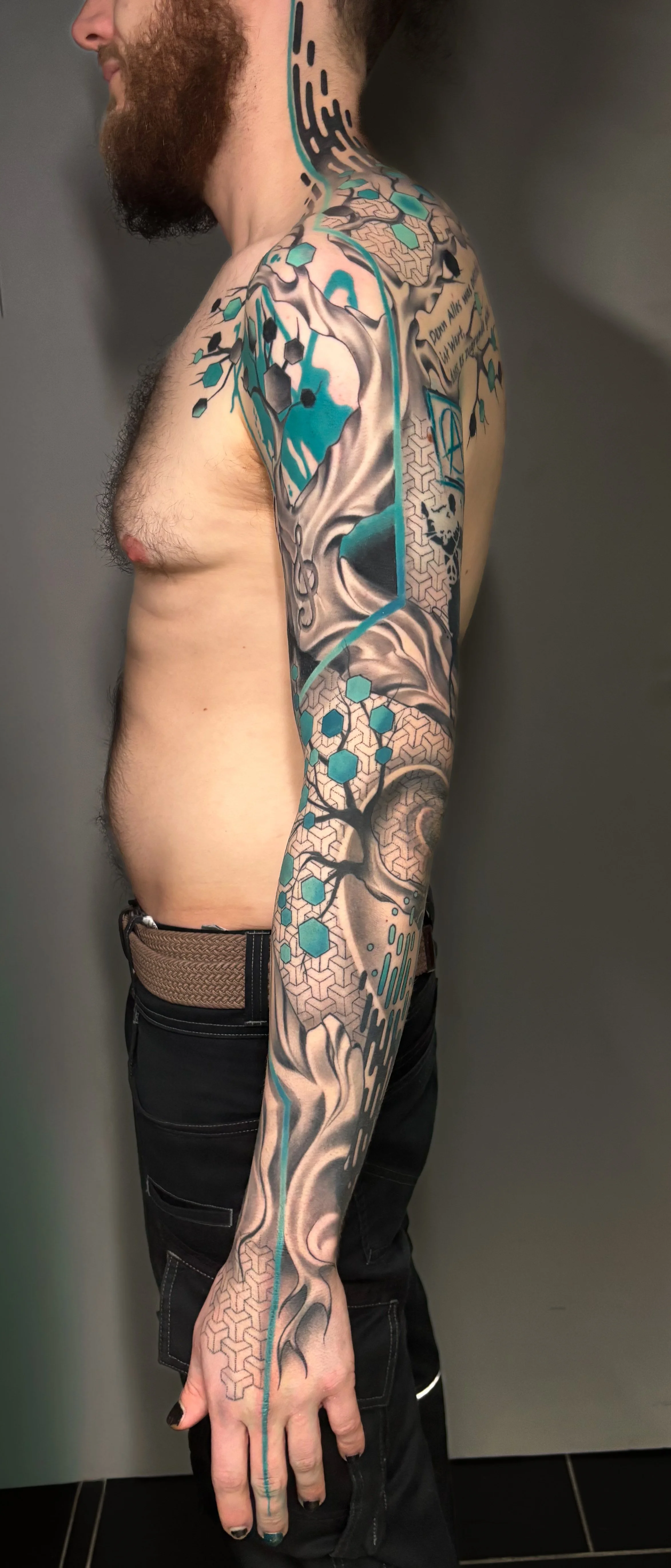 A man with a beard and tattoos on his arm, shoulder, chest, and neck, standing against a gray background. The tattoos feature abstract designs, geometric patterns, and stylized imagery with black, teal, and gray colors.