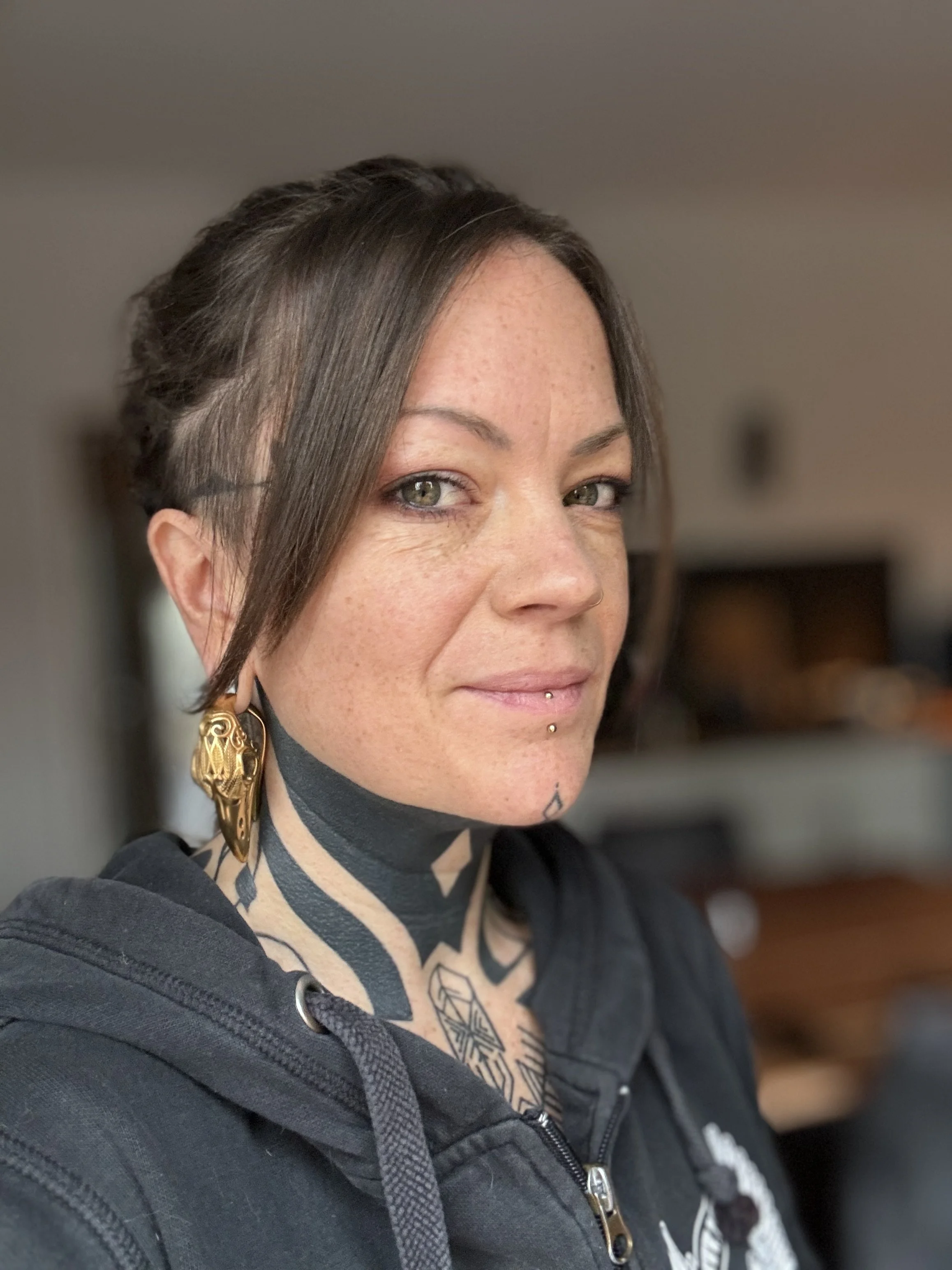 Close-up of a woman with light skin and gray eyes, wearing large gold earrings, a septum piercing, lip piercings, tattoos on her upper chest, and a dark hoodie, indoors.