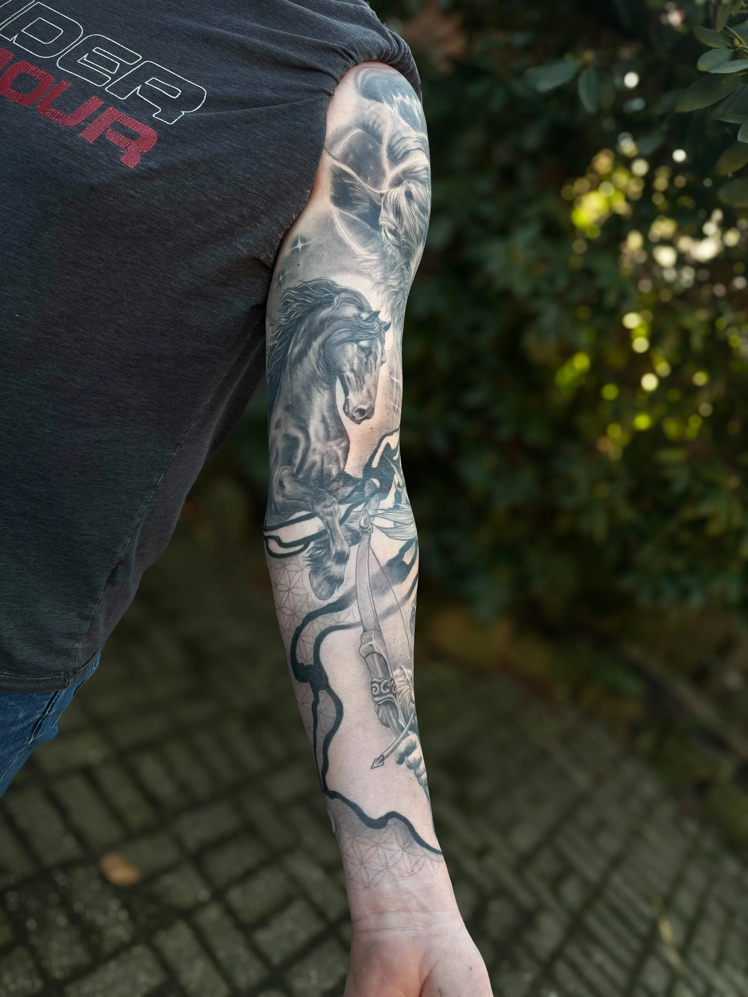 A person's arm with a detailed black and gray tattoo of a horse, a sword, and a star, against a background of green leaves and cobblestone ground.