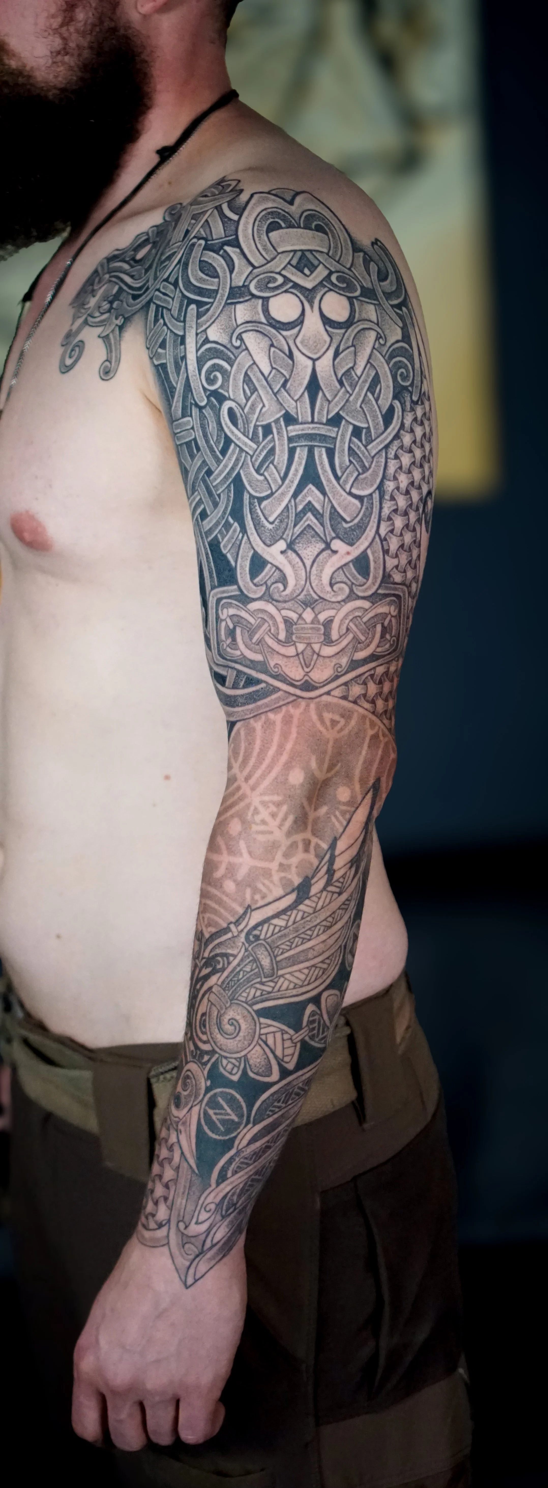Close-up of a man's upper arm and shoulder with intricate black and gray tattoos featuring Norse or Celtic knotwork, a face mask design, and Viking elements, wearing a black necklace and dark brown shorts.