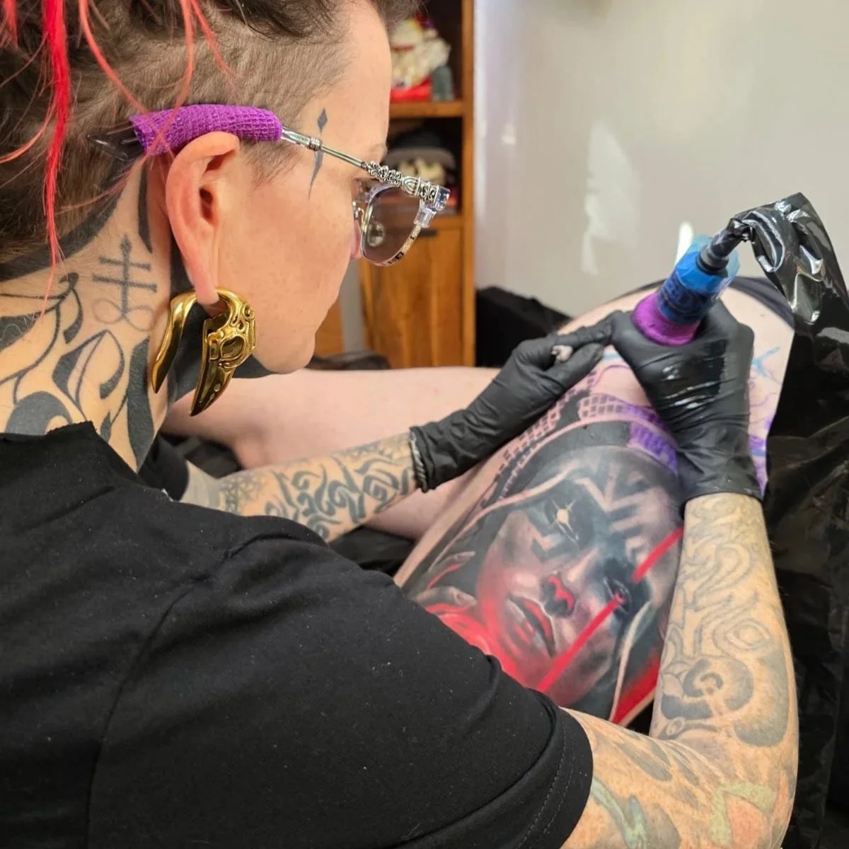 Tattoo artist working on a colorful portrait tattoo on a client's thigh with tattoo equipment, gloves, and safety glasses in a tattoo studio.