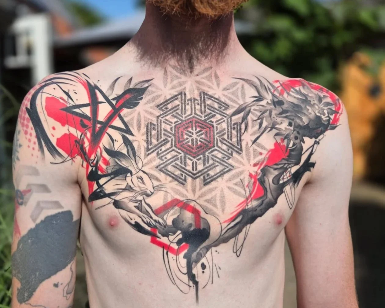 Tattooed man with a geometric and abstract tattoo design on his chest and arms, featuring a central hexagonal pattern, lions, and red accent splashes.