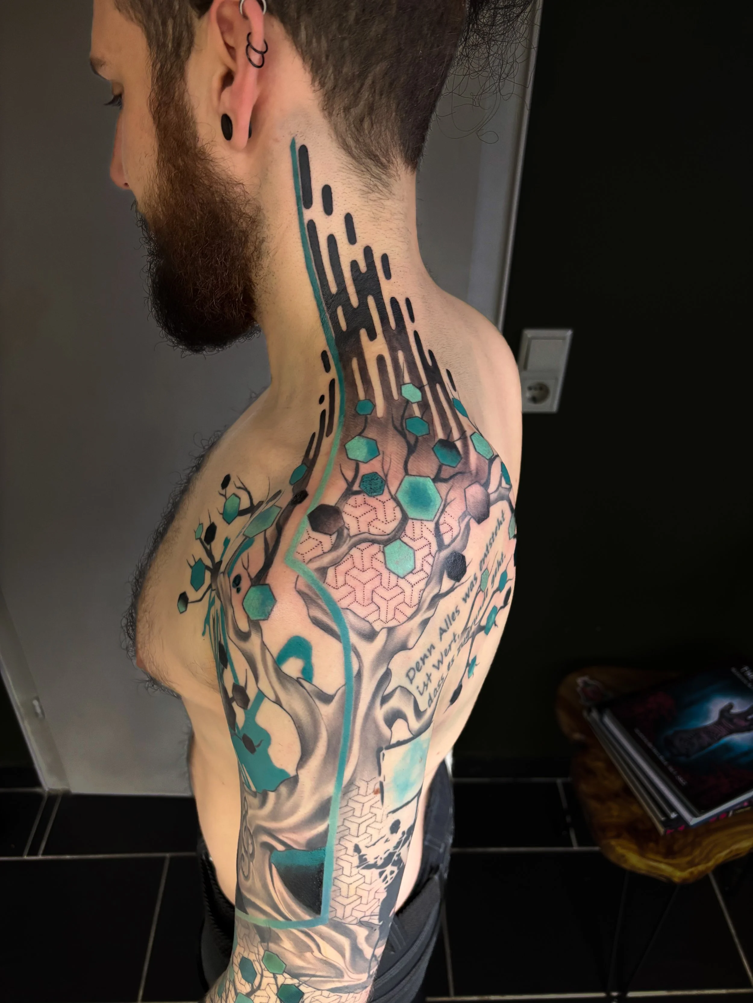 A man with tattoos on his shoulder and neck, featuring geometric and biological patterns, standing against a dark background in a room with black tile flooring and a shelf with books and a leather bag.