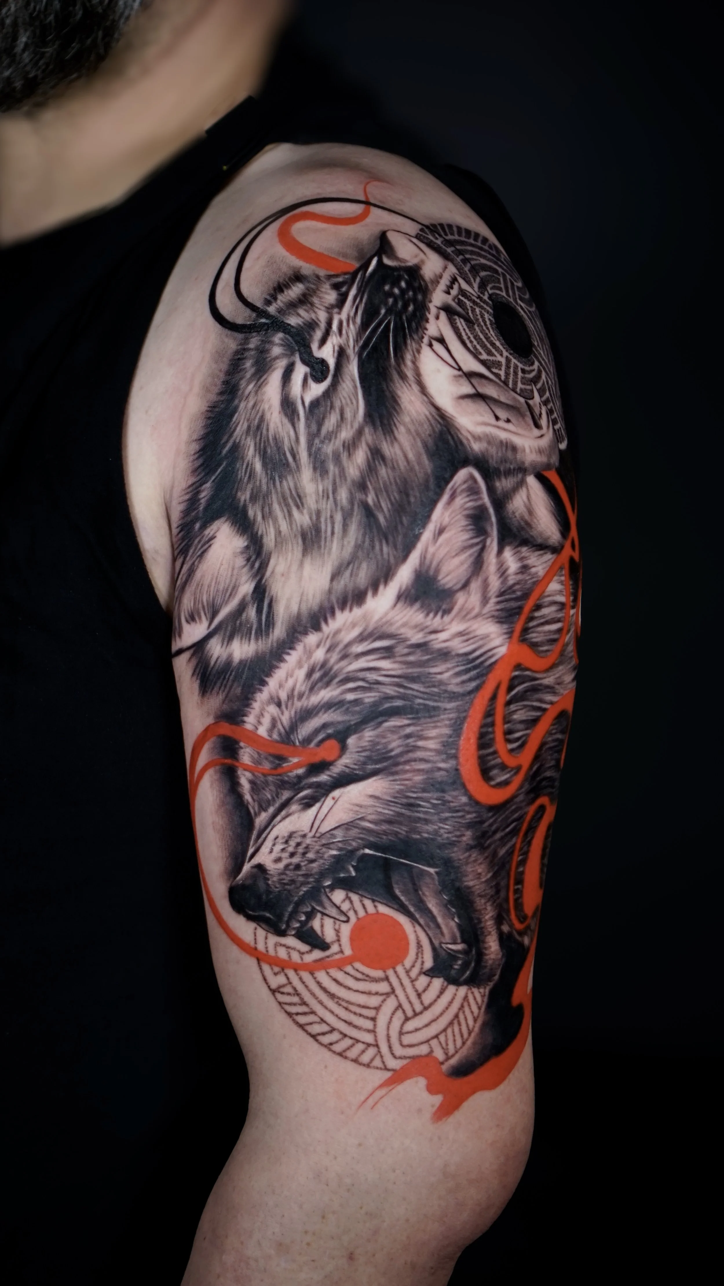 Tattoo of a wolf with detailed fur, snout, and open mouth, surrounded by abstract geometric designs and red accents on a person's upper arm.