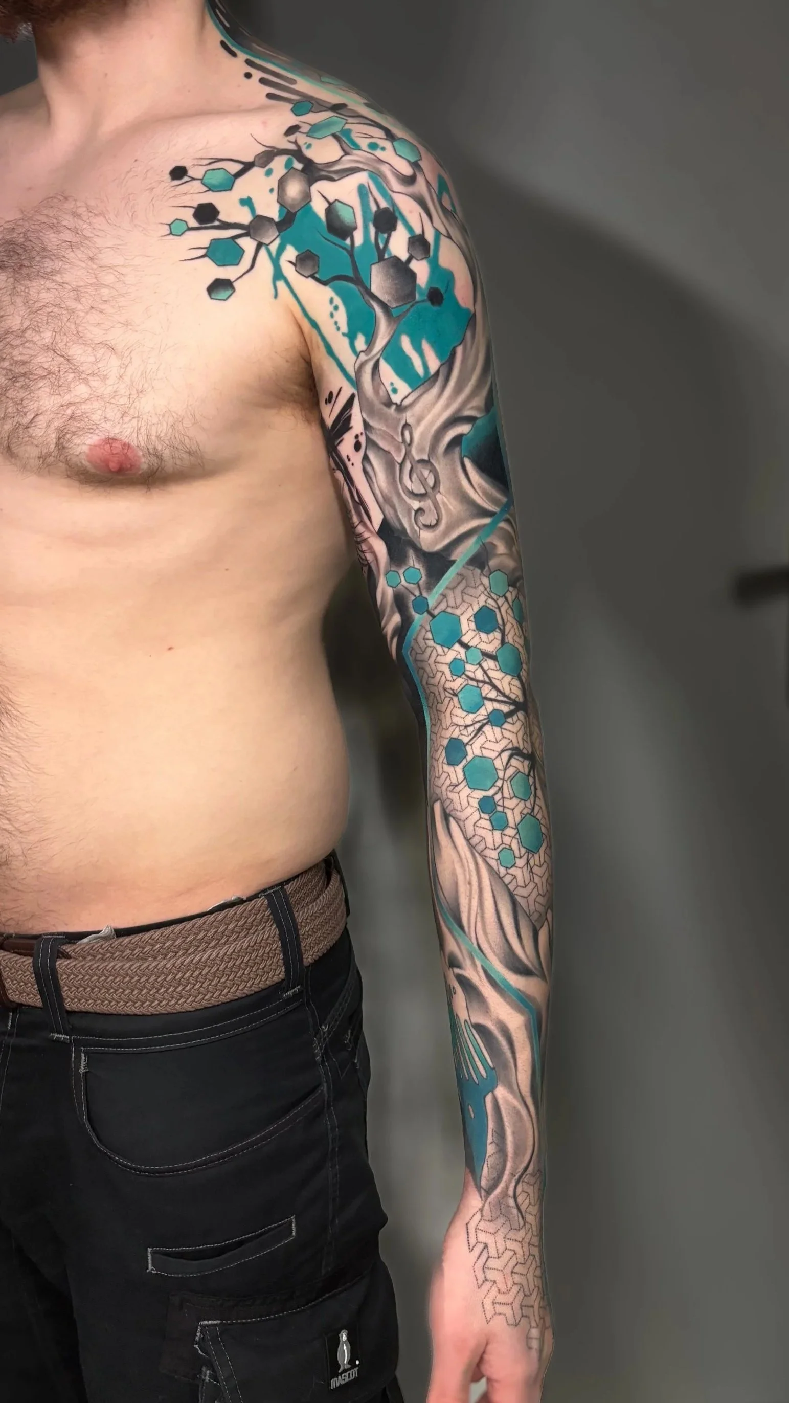 A person with a detailed arm tattoo featuring a tree, molecular structures, and hexagonal patterns in black, gray, teal, and blue colors.