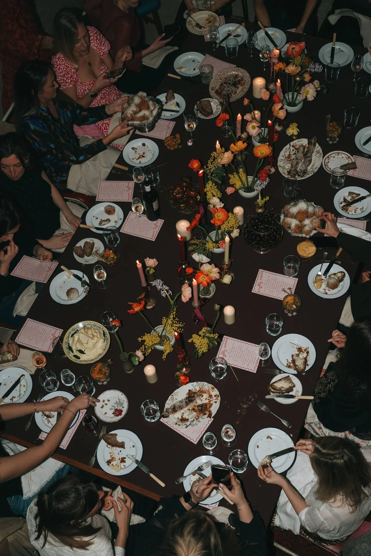 A group of women gathered around a large dinner table celebrating, with empty dessert plates, glasses of wine, and floral centerpieces with lit candles.