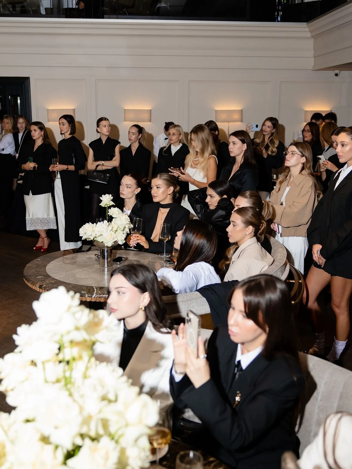 A group of women at a formal event, some standing and some sitting, dressed in elegant black, beige, and white outfits, with glasses of wine and floral centerpieces on the table.