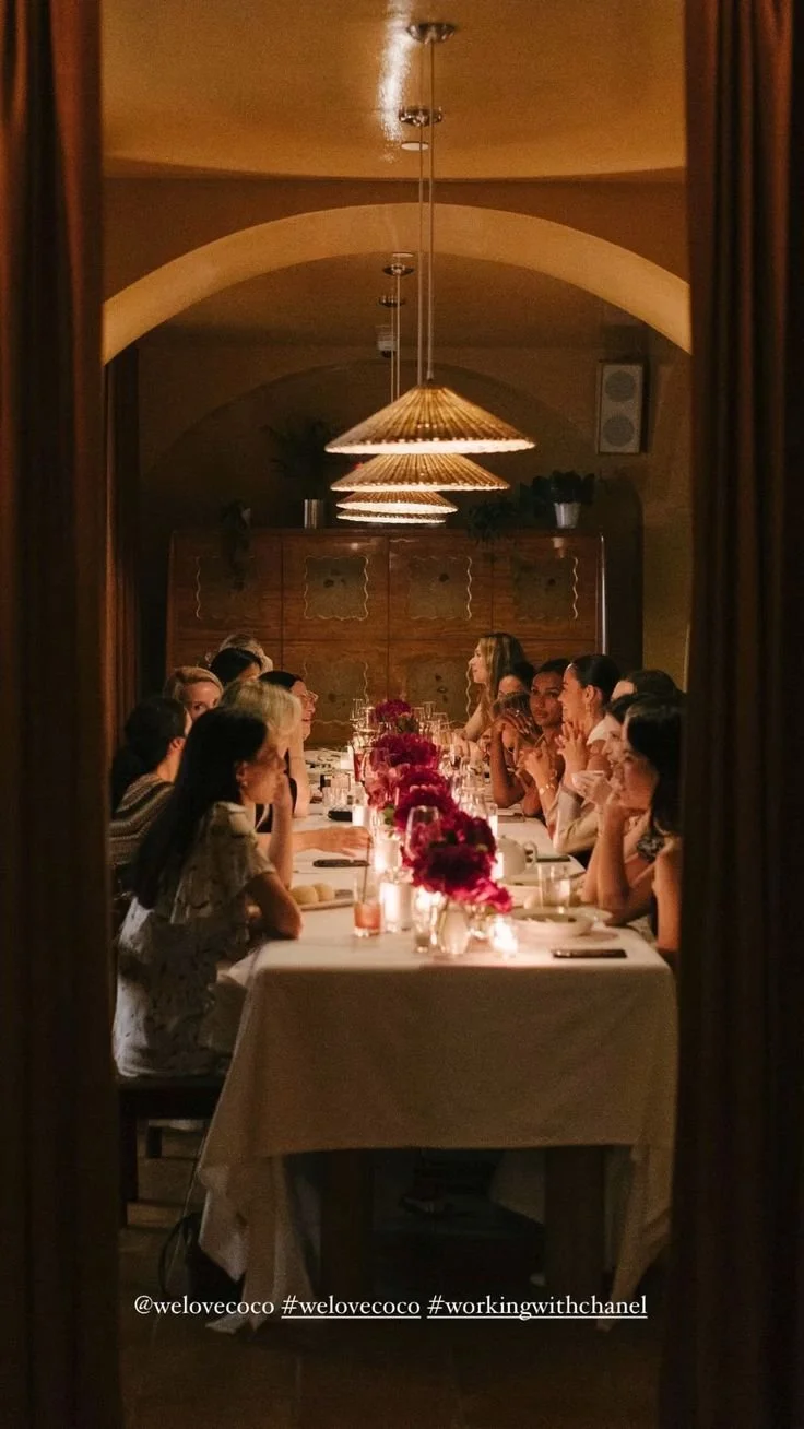 People sitting at a long dinner table with pink flowers and candles, seen through a doorway, in a warmly lit restaurant or banquet hall.
