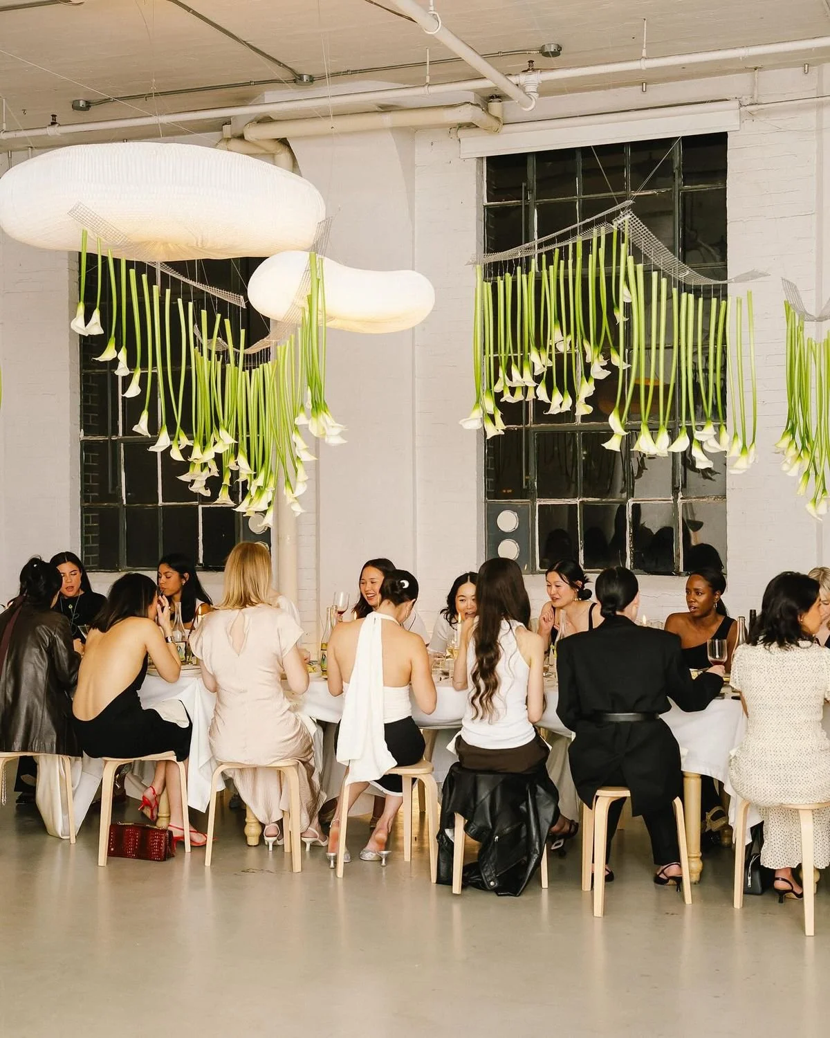 People dining at a long table in a decorated, modern indoor space with large windows and hanging floral and cloud-like installations.