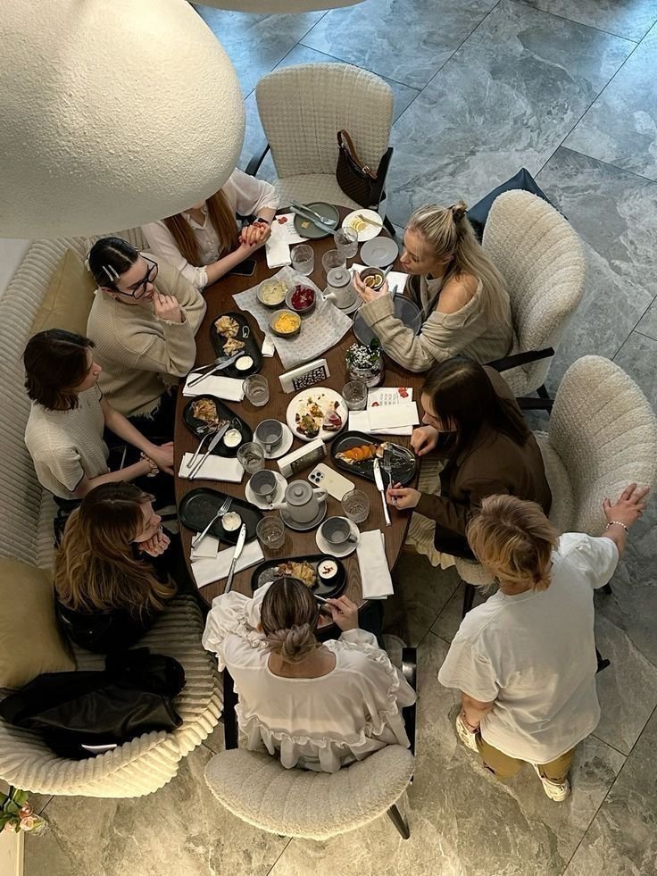Top-down view of a group of nine women gathered around a dining table, enjoying a meal with various dishes, drinks, utensils, and notebooks, in a restaurant setting with marble flooring.