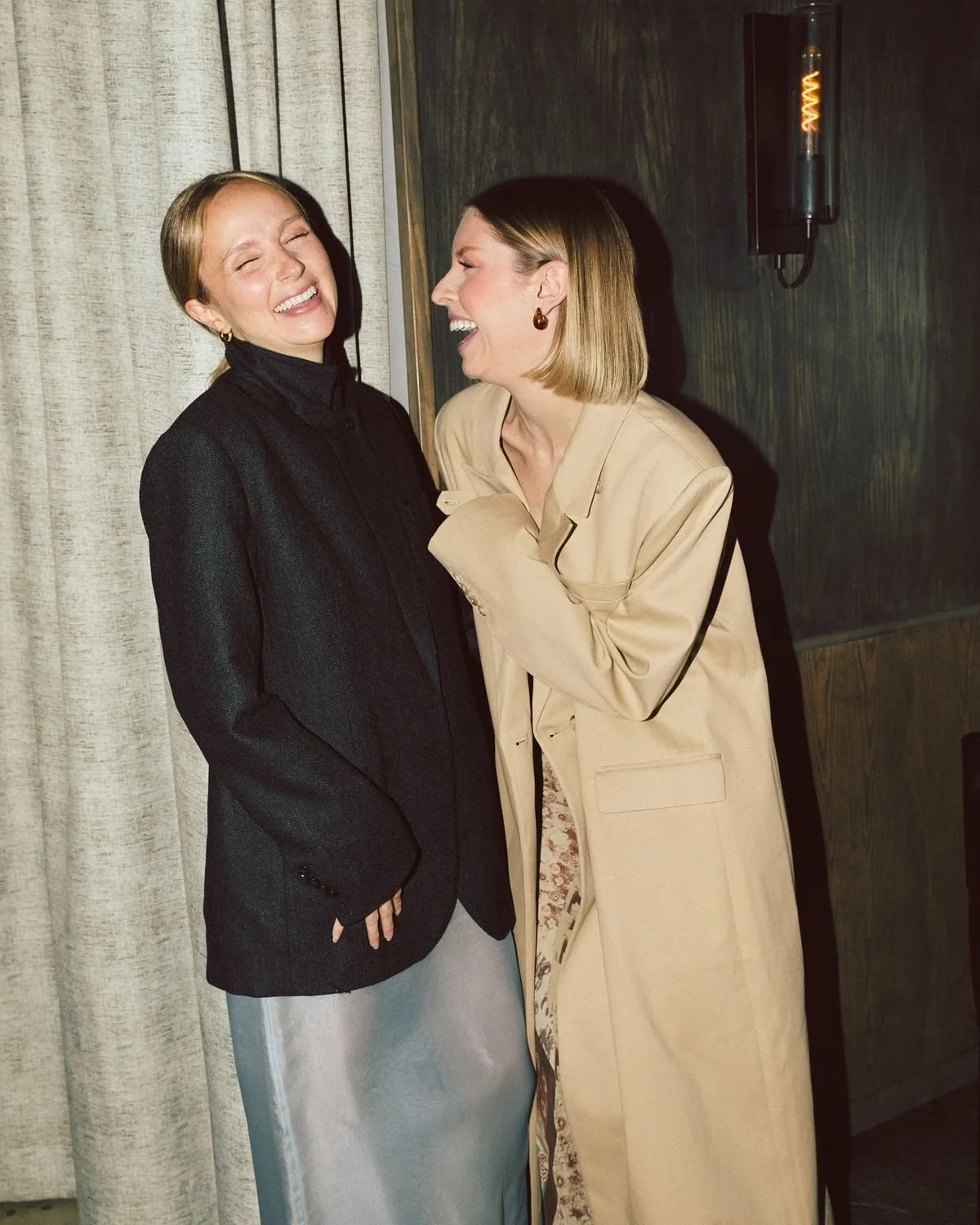Two women laughing and enjoying each other's company indoors. One woman is wearing a black blazer and gray skirt, and the other woman is wearing a beige coat. They are standing close to each other in front of a curtain and a wall with a light fixture.