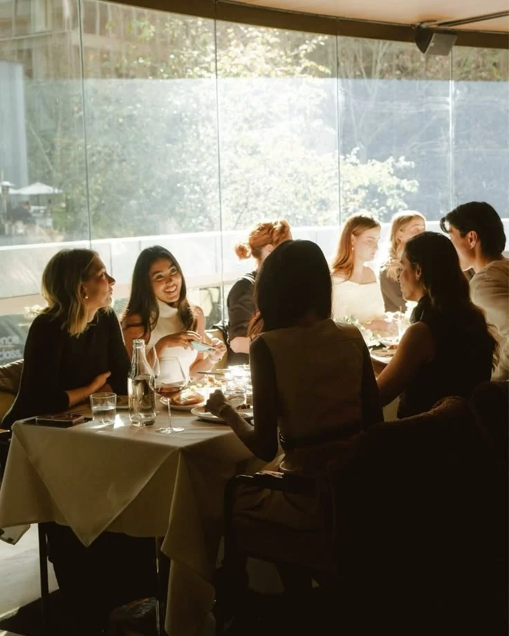 A group of women and one man sitting around a dining table in a restaurant, engaged in conversation, with sunlight streaming in through large windows in the background.