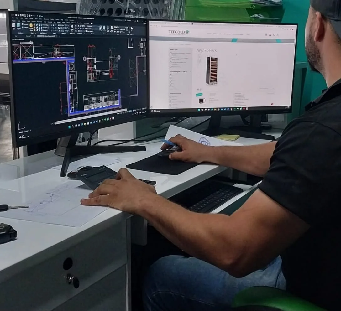 Persona trabajando en una estación de dos monitores, uno con un software de diseño técnico y otro con una página web de comercio, en un escritorio con papeles y herramientas.