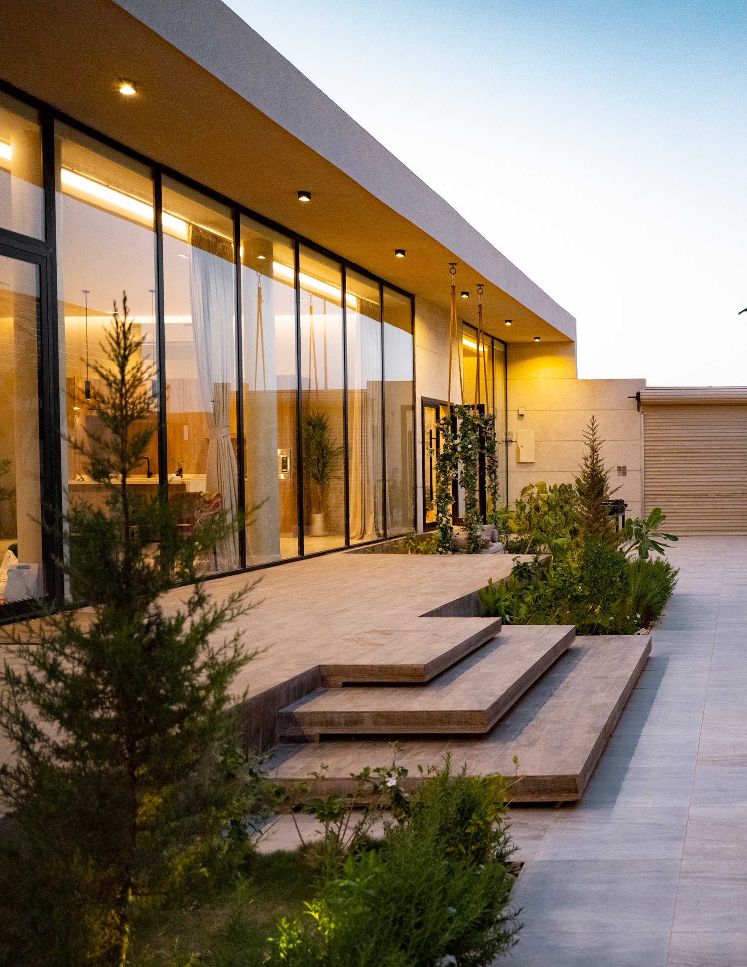 Modern house exterior with large glass windows, illuminated interior, outdoor plants, and stone steps at dusk.