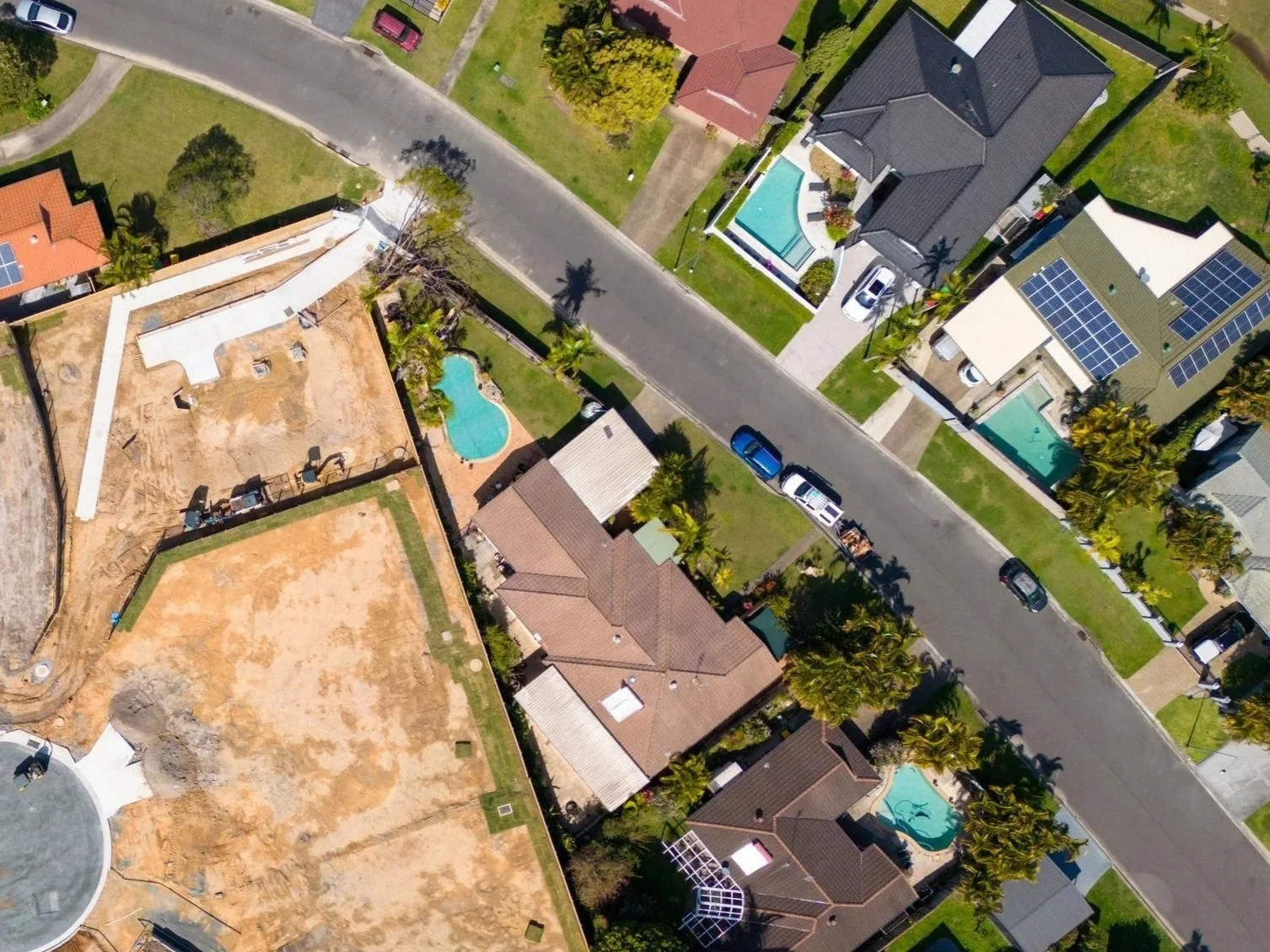 Aerial view of a residential neighborhood with houses, swimming pools, and a street with parked cars. Some houses have solar panels on their roofs, and there is a construction site with exposed ground and construction equipment.