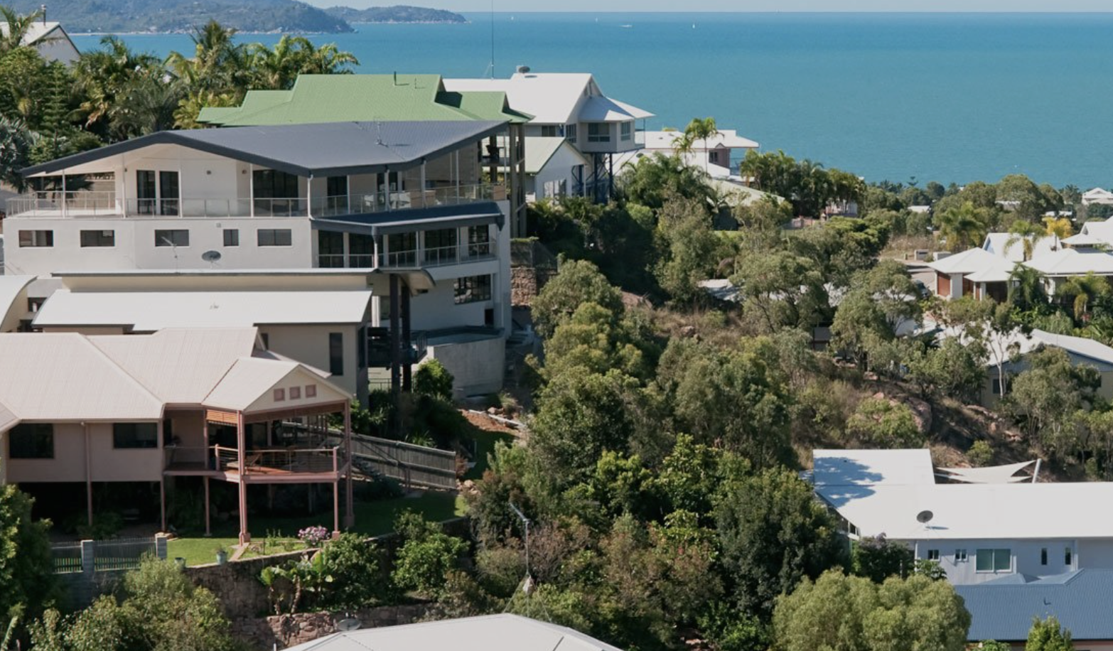 Aerial view of houses on a hillside near the ocean with lush green trees and clear blue water in the background.