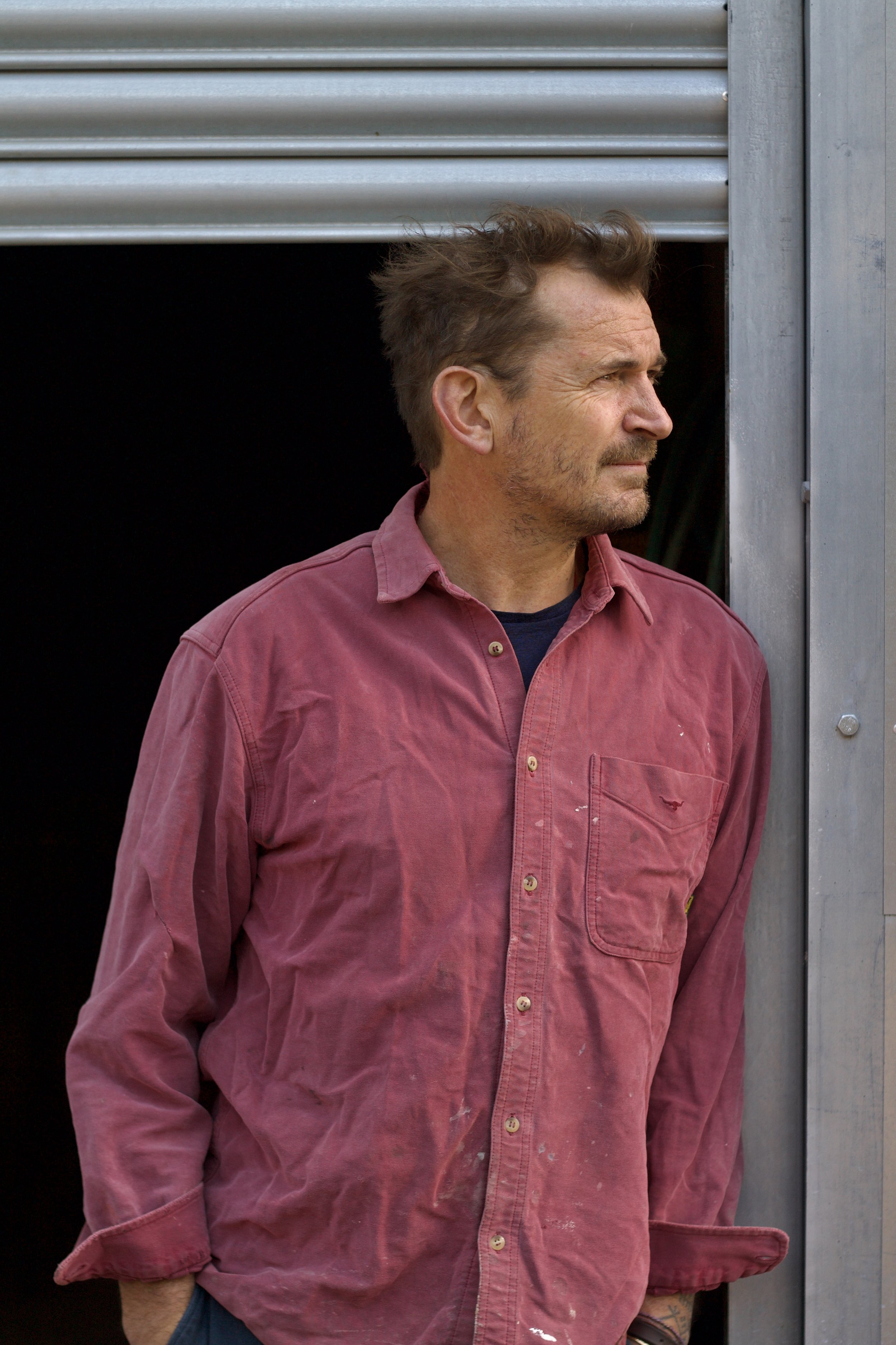 A man with brown hair and a beard wearing a red button-up shirt stands outside, looking to his right, near a metal structure.
