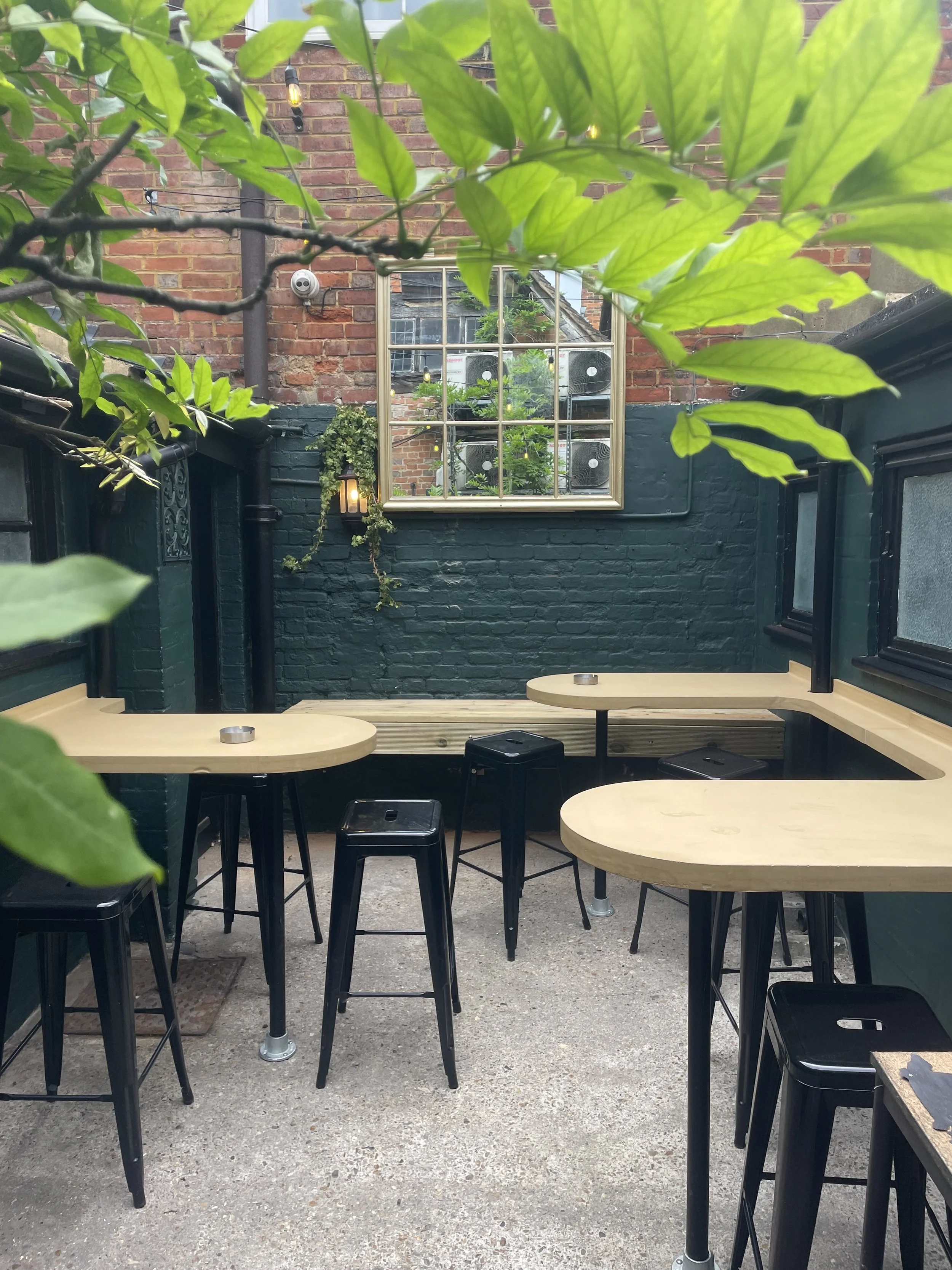 Outdoor seating area with light-colored wooden tables and black stools, against a dark green painted brick wall with a window and some plants, in an urban patio setting.