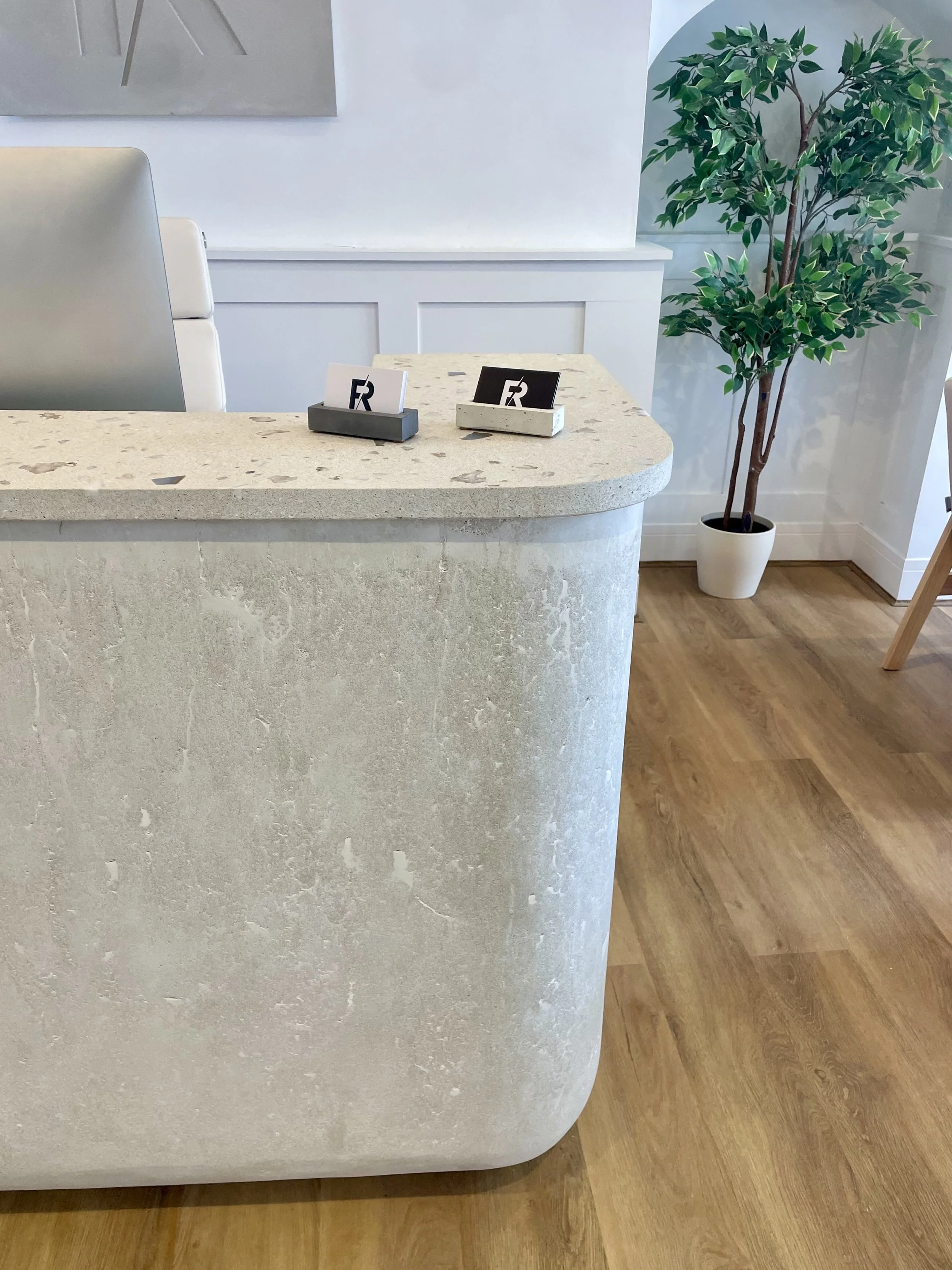 A reception area with a curved, light-colored stone counter, two small menu or brochure holders, a white wall, a large potted green plant, light wood flooring, and a white chair.