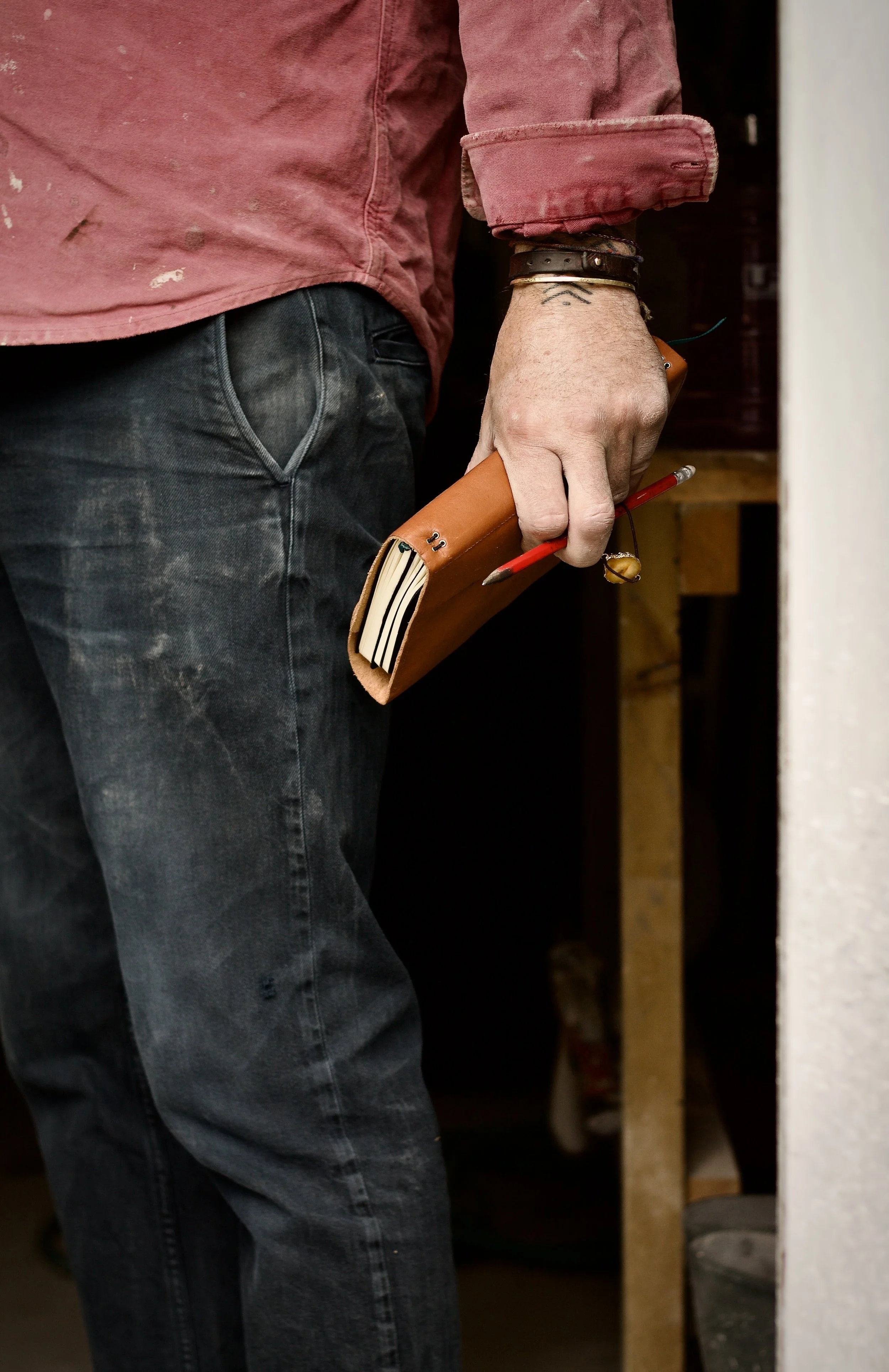 Person wearing black jeans and a red, paint-splattered long-sleeve shirt, holding a brown leather notebook with papers inside and a red pen, in a workshop or studio setting.