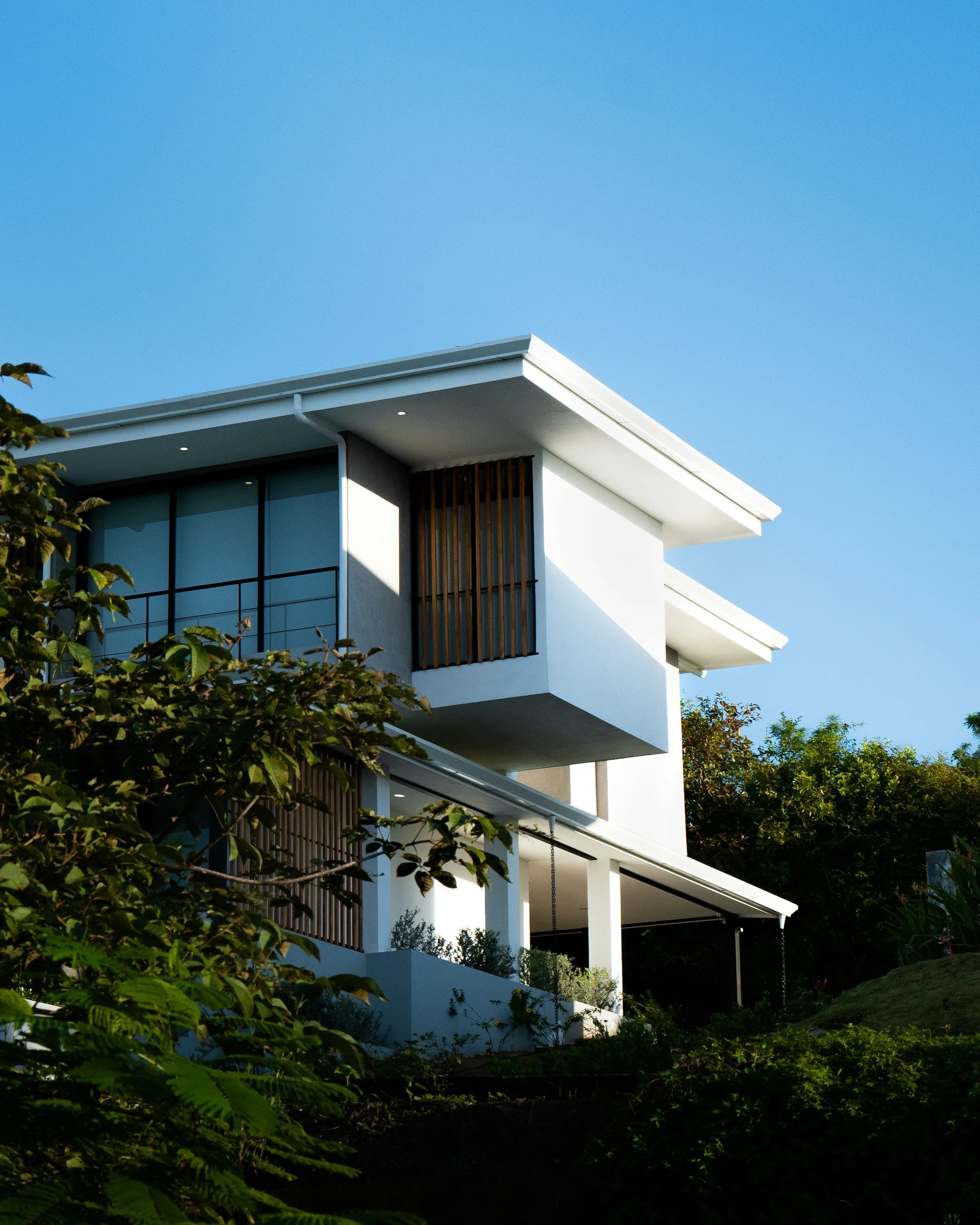 Modern white multi-story house with large windows and wooden accents, surrounded by greenery, under a clear blue sky.