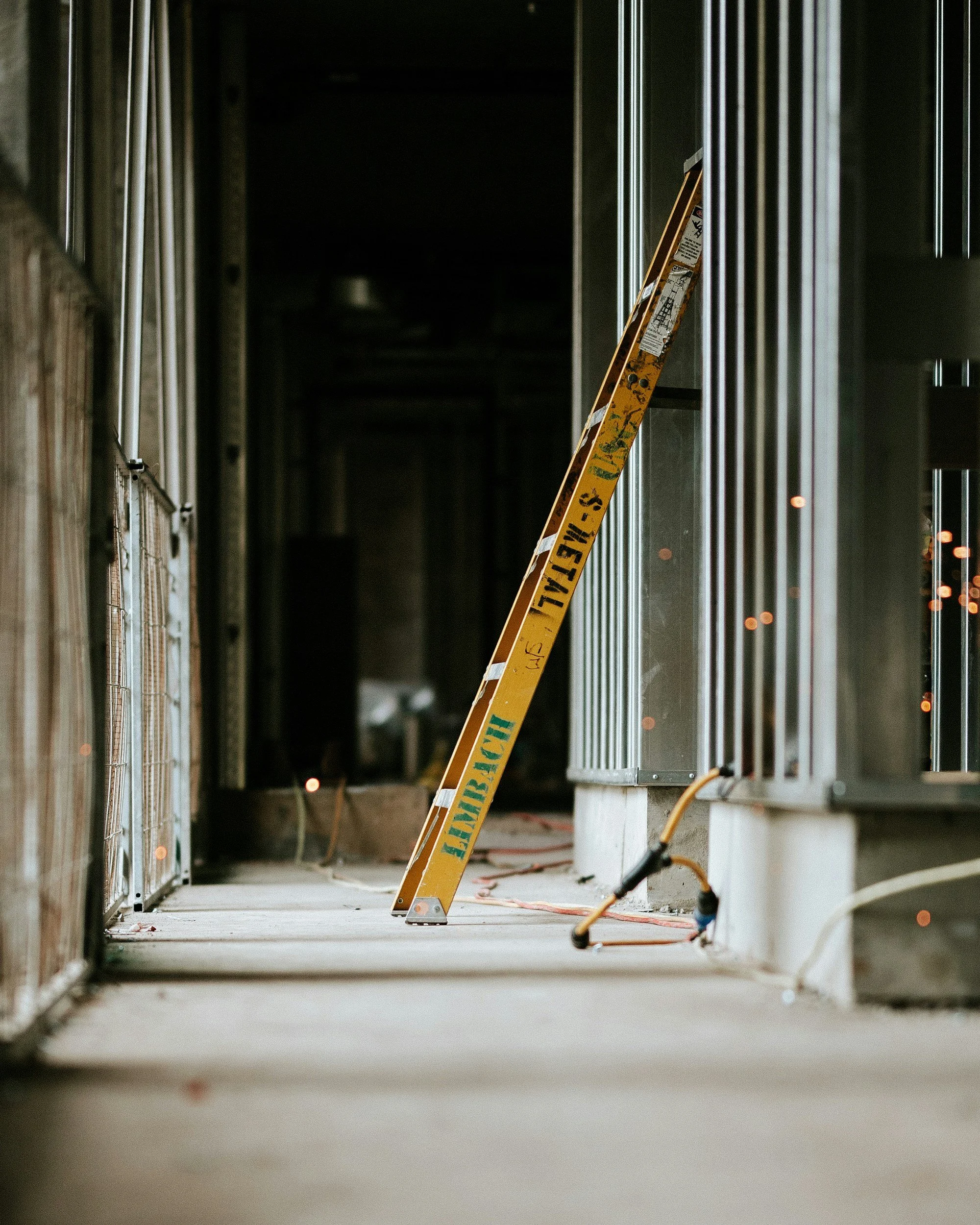 A construction site hallway with exposed framing and wiring, featuring a yellow safety ladder leaning against the wall, with caution tape on it.